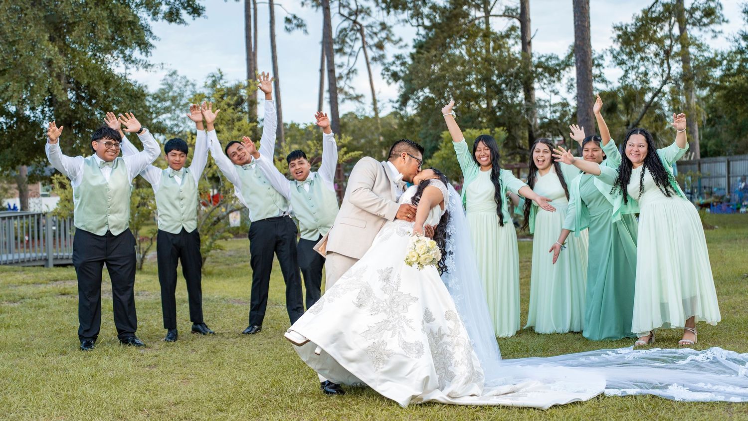 Joyful wedding party celebrating with raised arms as groom dips bride outdoors on sunny day.