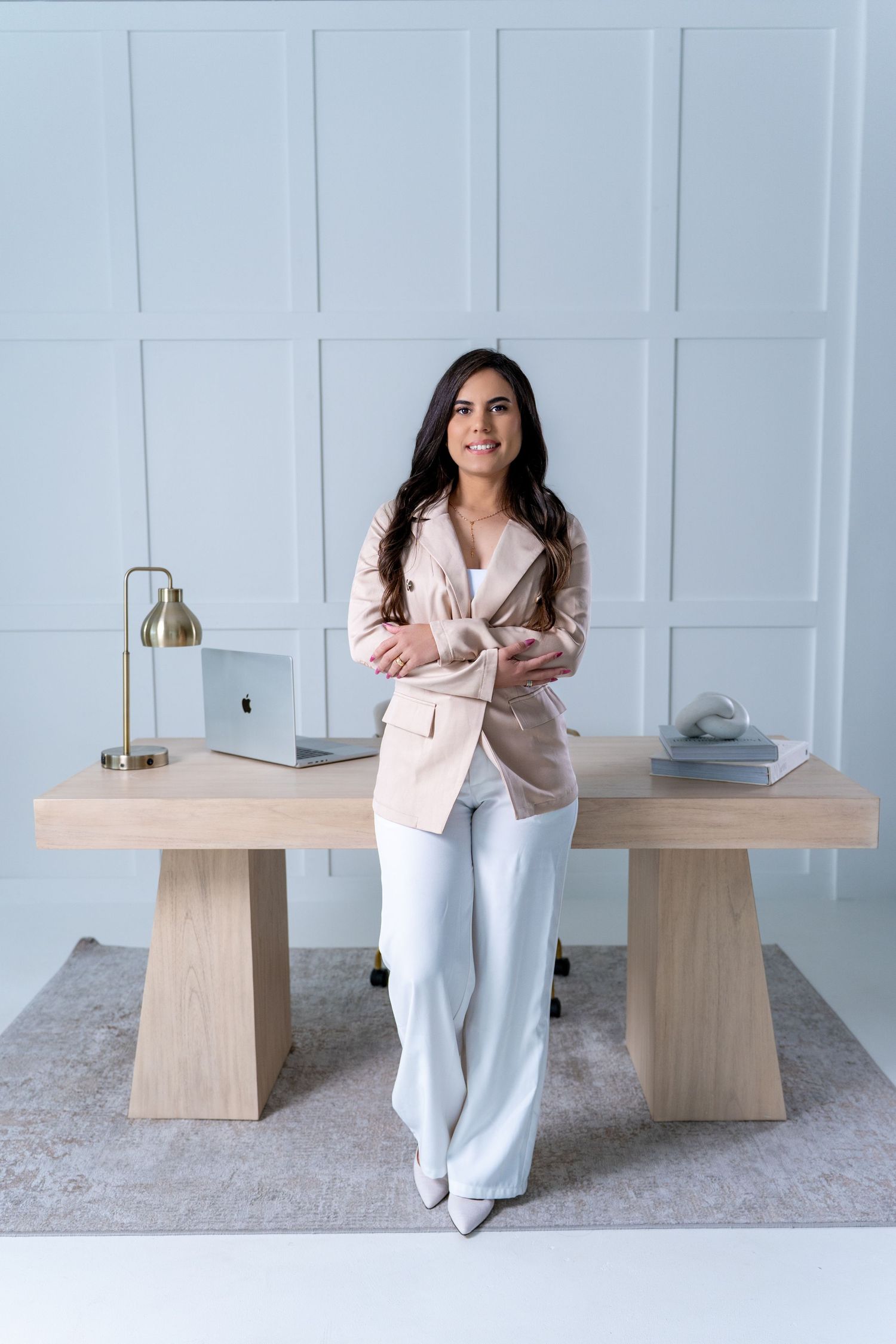 Professional woman in beige blazer and white pants stands confidently by modern desk with laptop in minimalist office setting.