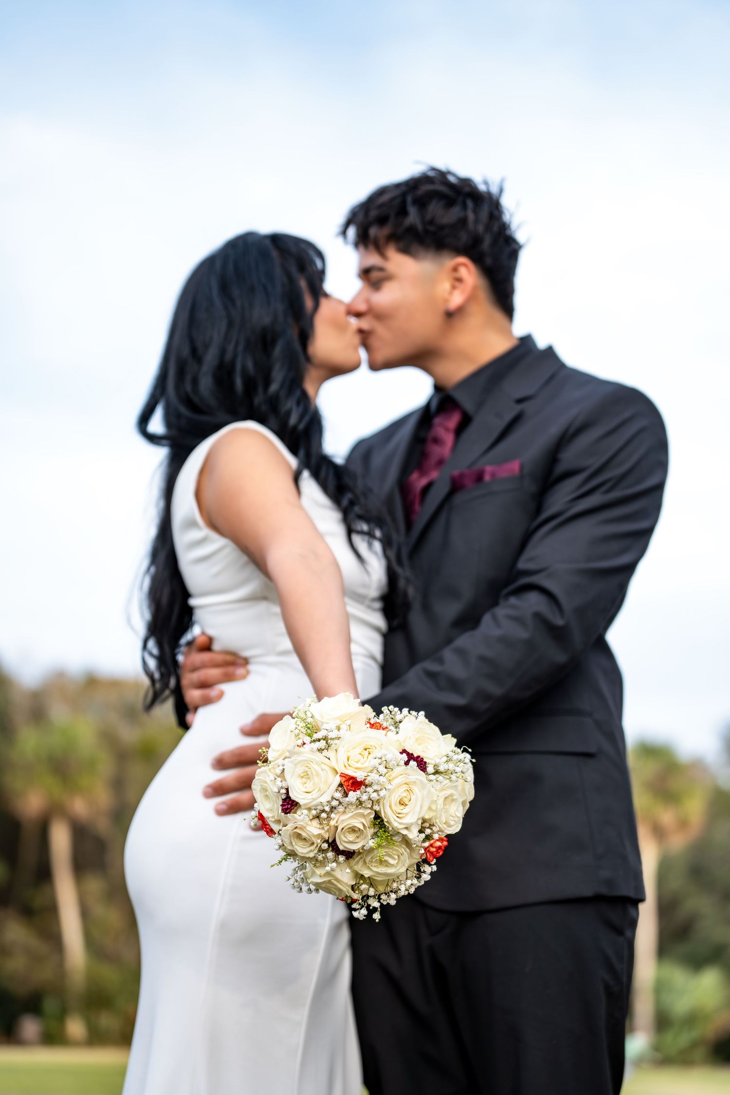 A couple shares an intimate moment against a nature backdrop with a white floral bouquet.