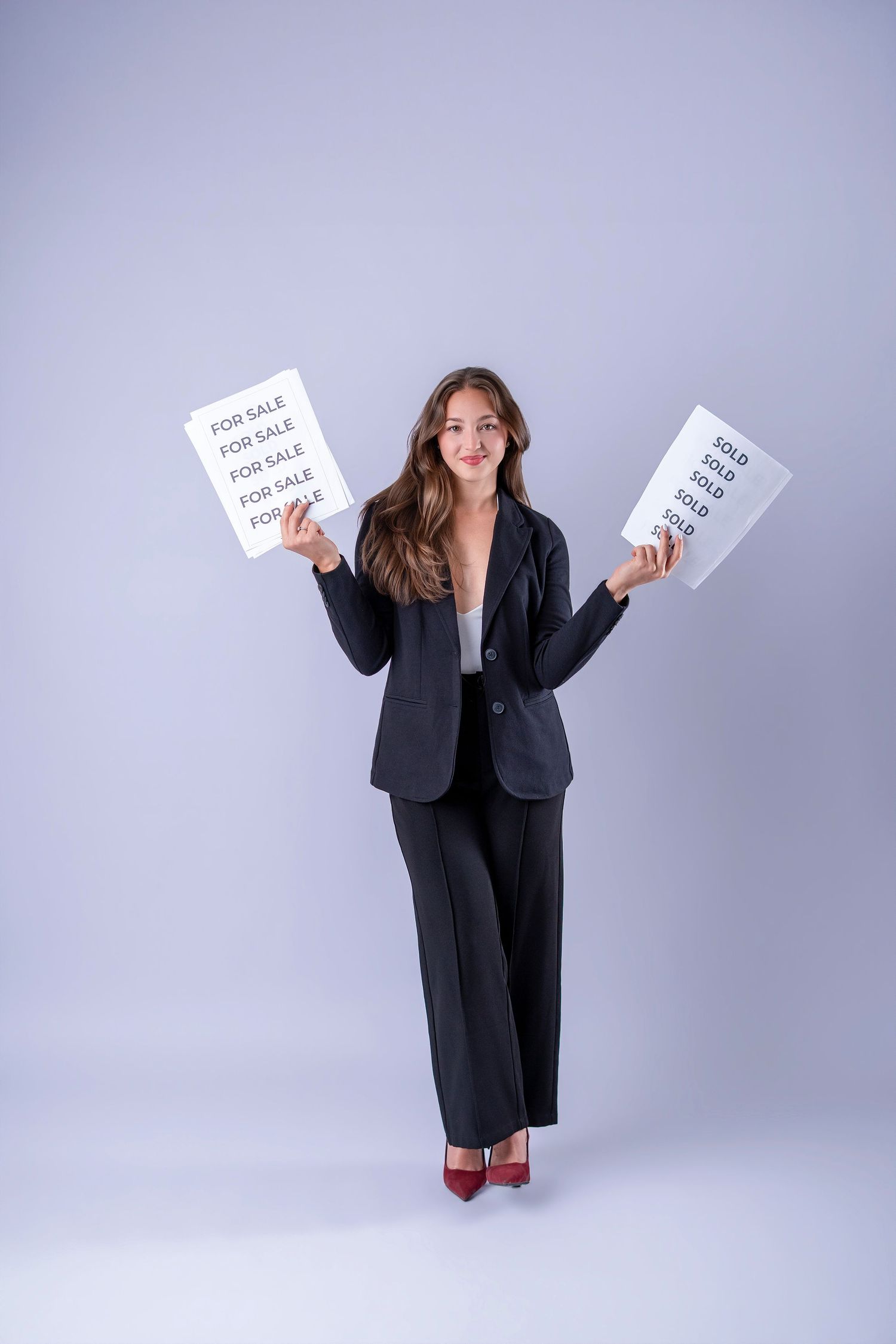 Professional female real estate agent in black suit holding 'For Sale' and 'Sold' signs on gray background.