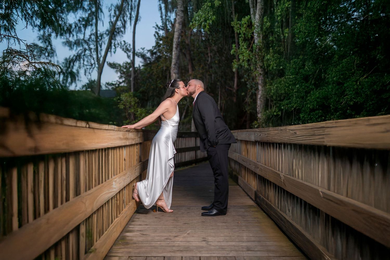 Bride and groom sharing a romantic kiss on a wooden bridge surrounded by lush green trees on their wedding day.
