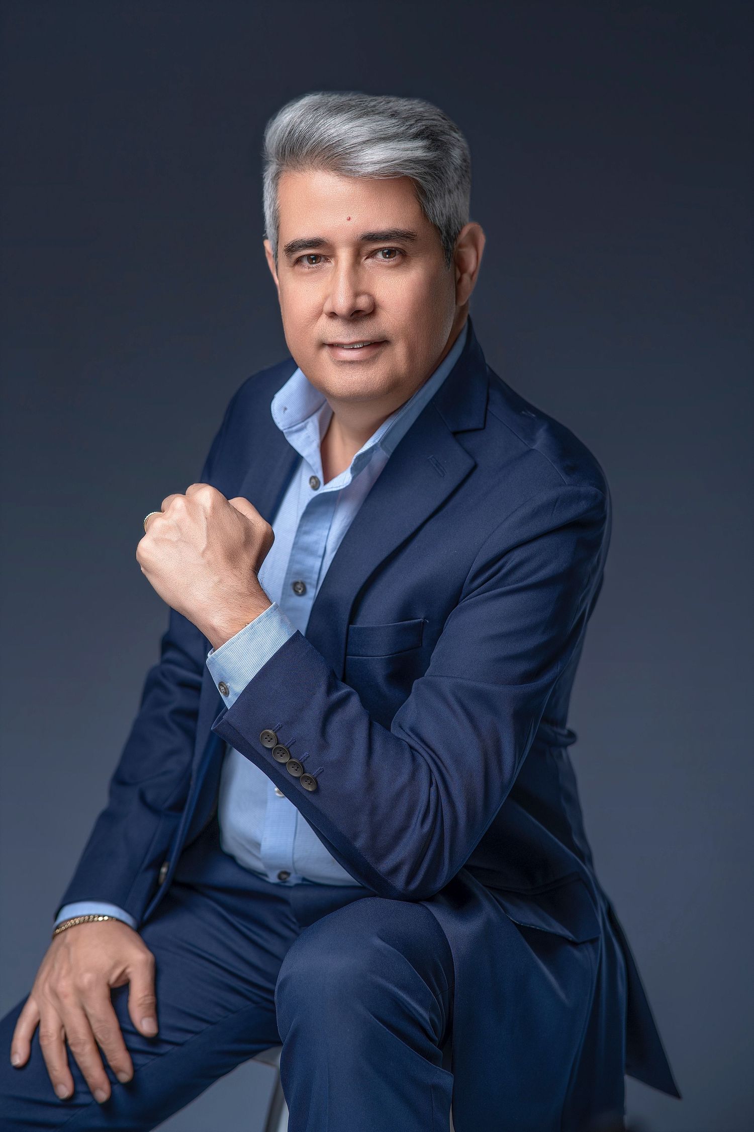 Middle-aged man with gray hair in navy blue suit sitting confidently with raised fist on gray background.