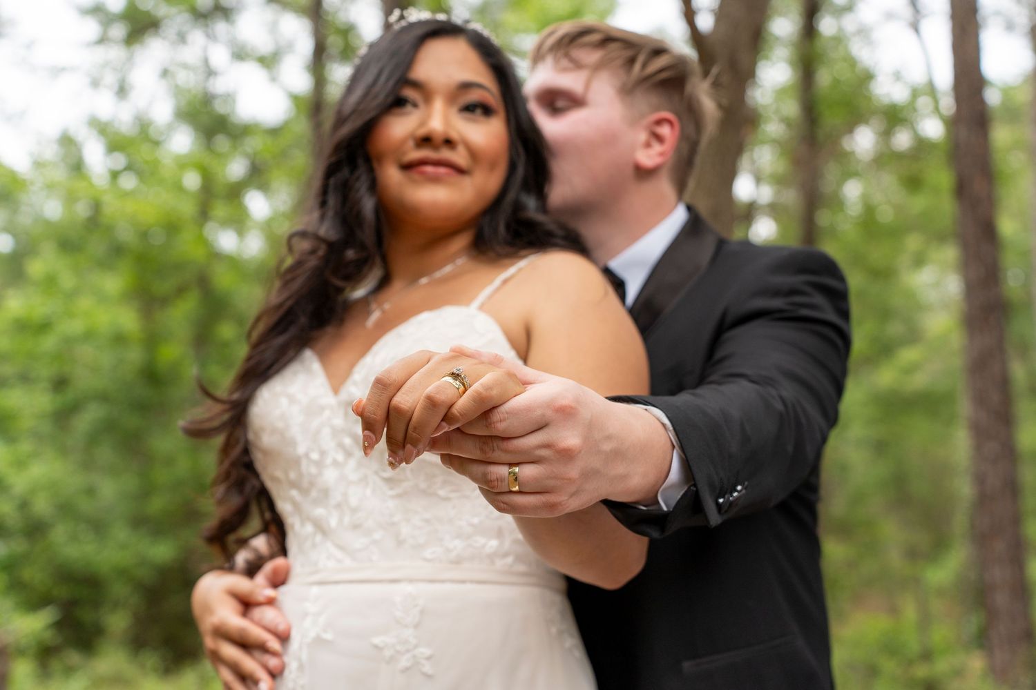 Bride and groom hold hands in a forest, wearing wedding attire, as groom kisses bride on the cheek.