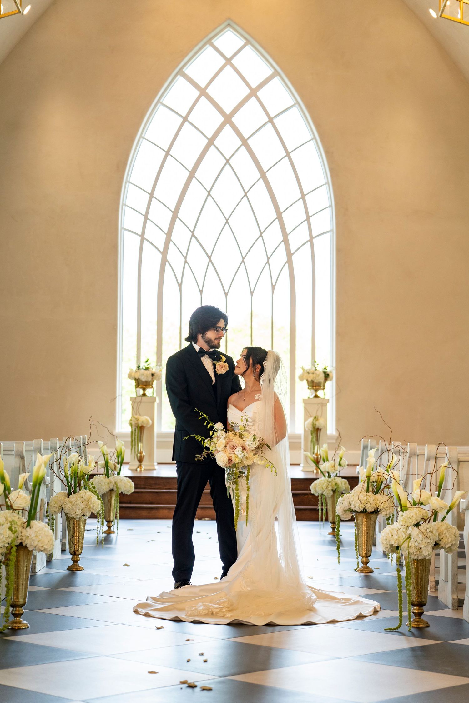 Bride and groom portrait inside chapel with gothic arch window and floral aisle.