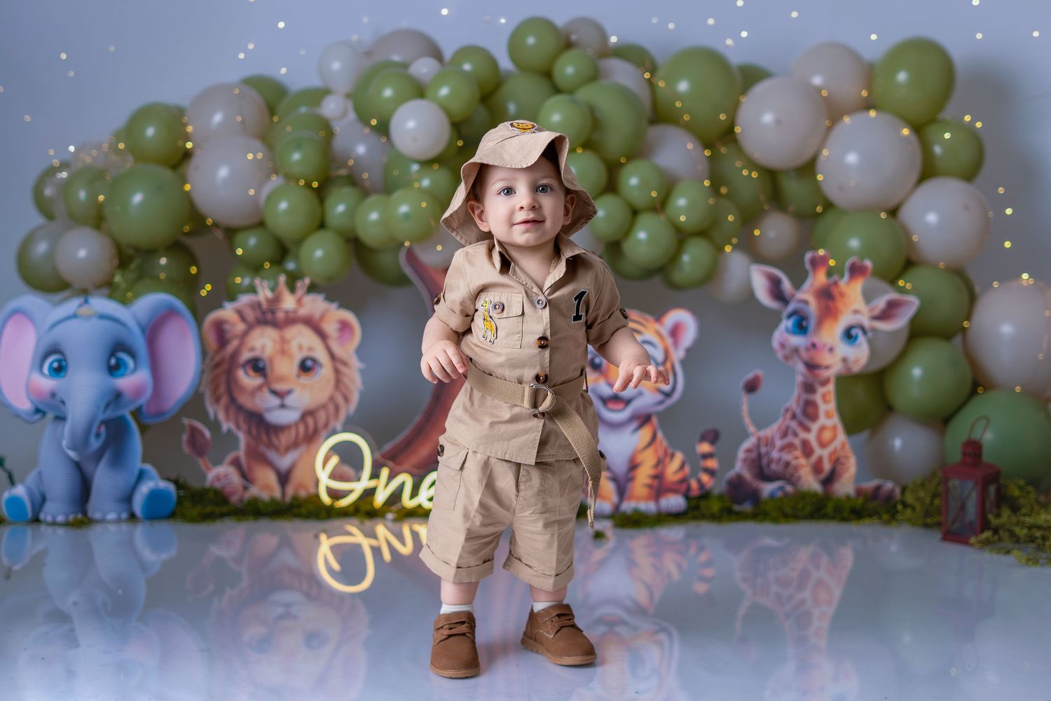 Toddler in safari outfit poses before balloon arch with jungle animal decorations at a first birthday party.