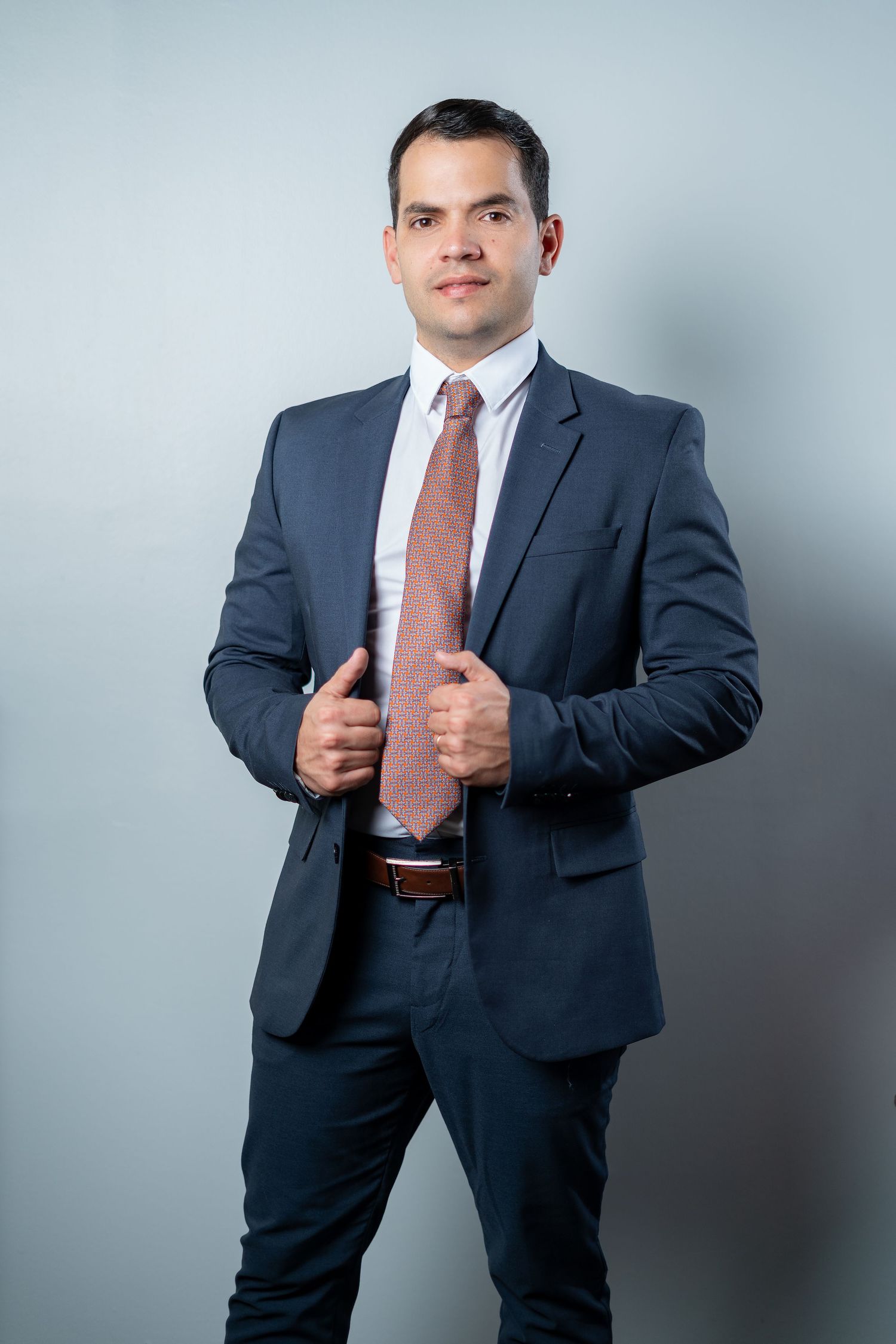 Professional wearing a navy suit and salmon tie adjusts his jacket while posing against a gray background.