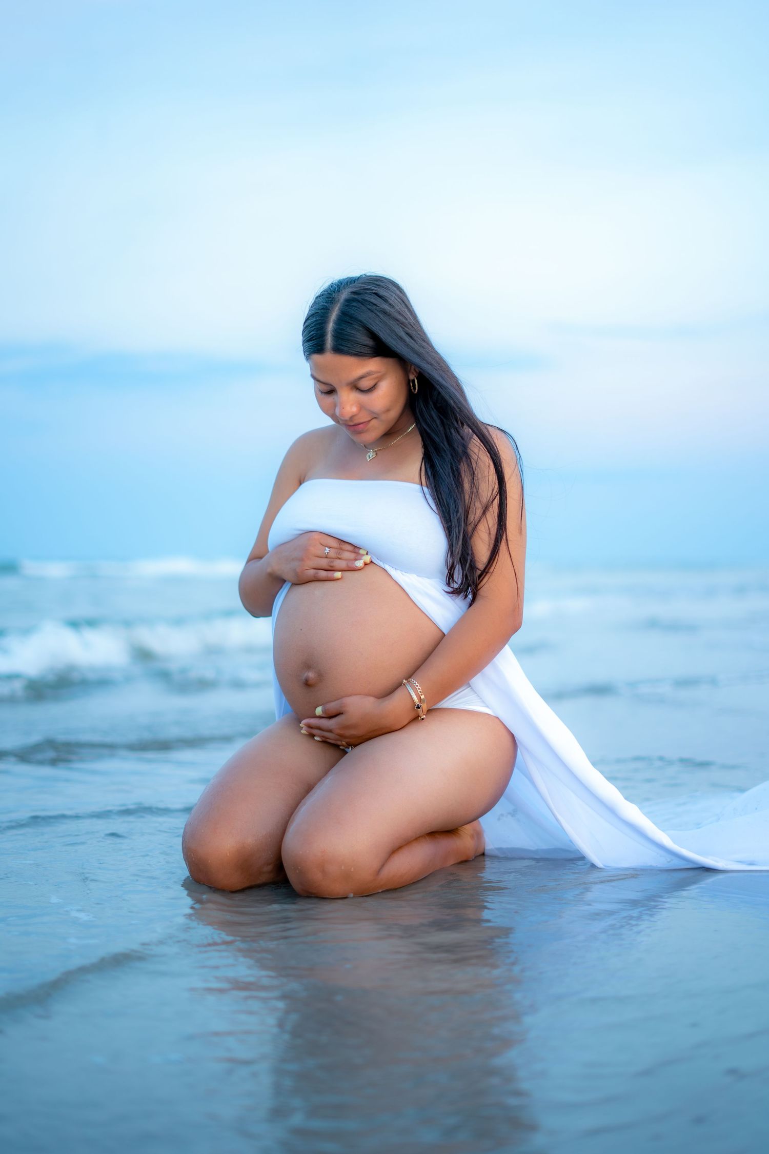 A silhouetted figure in white clothing sits peacefully at the ocean's edge during a serene beach maternity photoshoot.