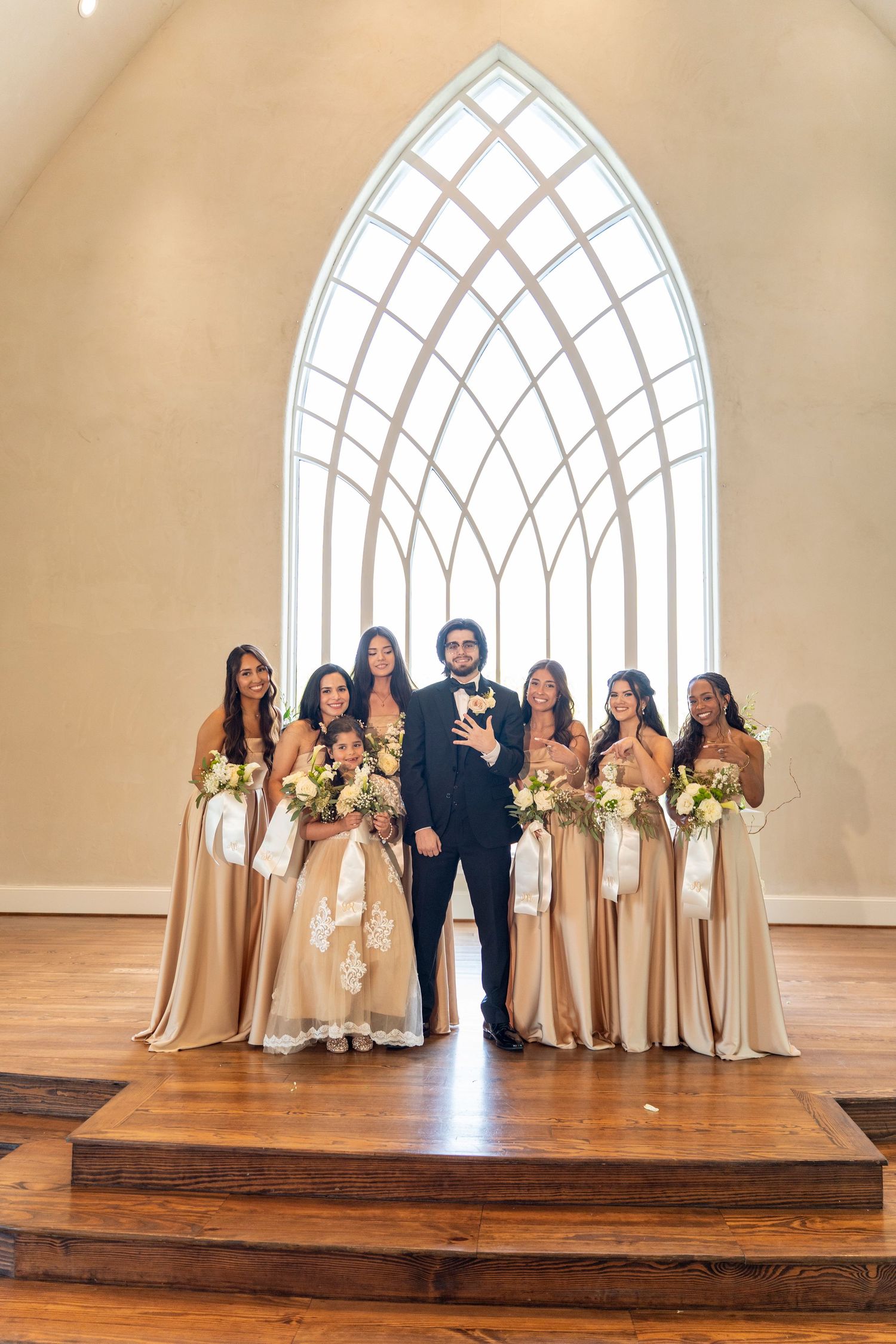 Groom in black tuxedo poses with bridesmaids in champagne gowns holding bouquets in elegant chapel.