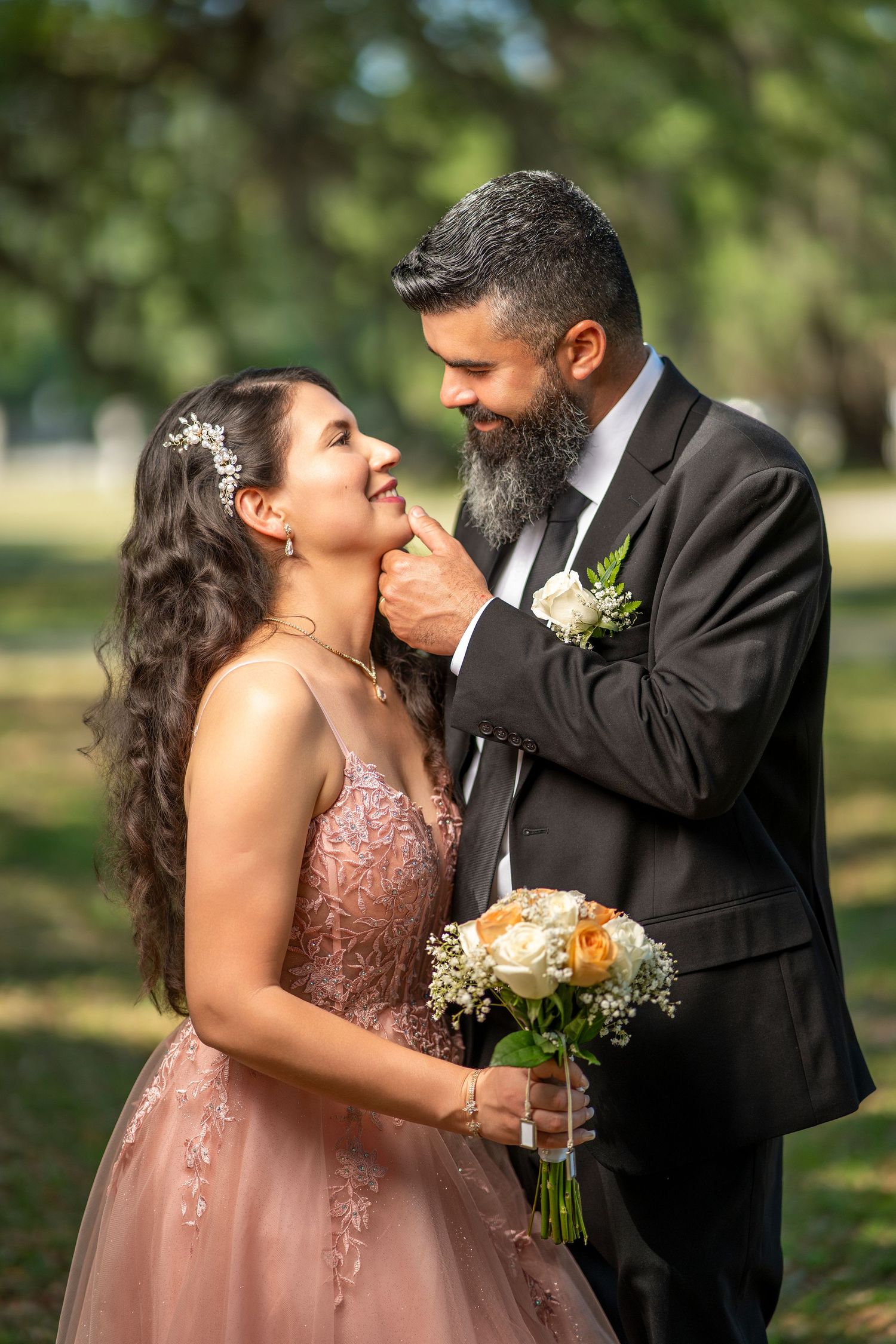 Smiling bride and bearded groom share tender moment outdoors, bride holding peach and white bouquet.