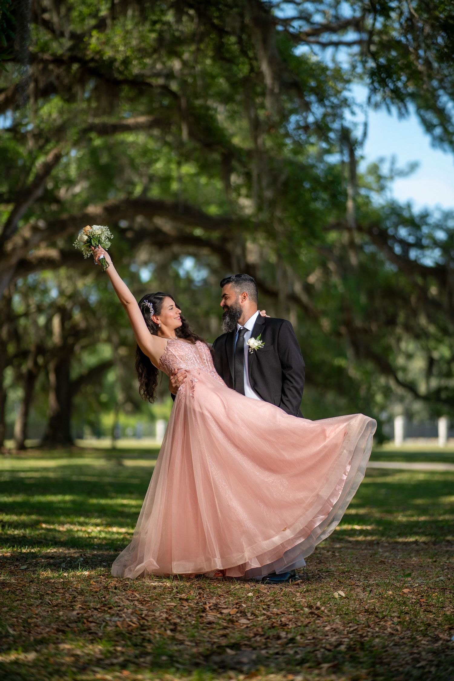 Bride in pink gown twirls bouquet while groom in black suit smiles under moss-draped oak trees.
