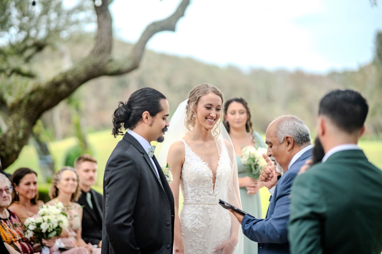 A laughter-filled moment captured at an outdoor wedding ceremony under trees with the wedding party in formal attire.