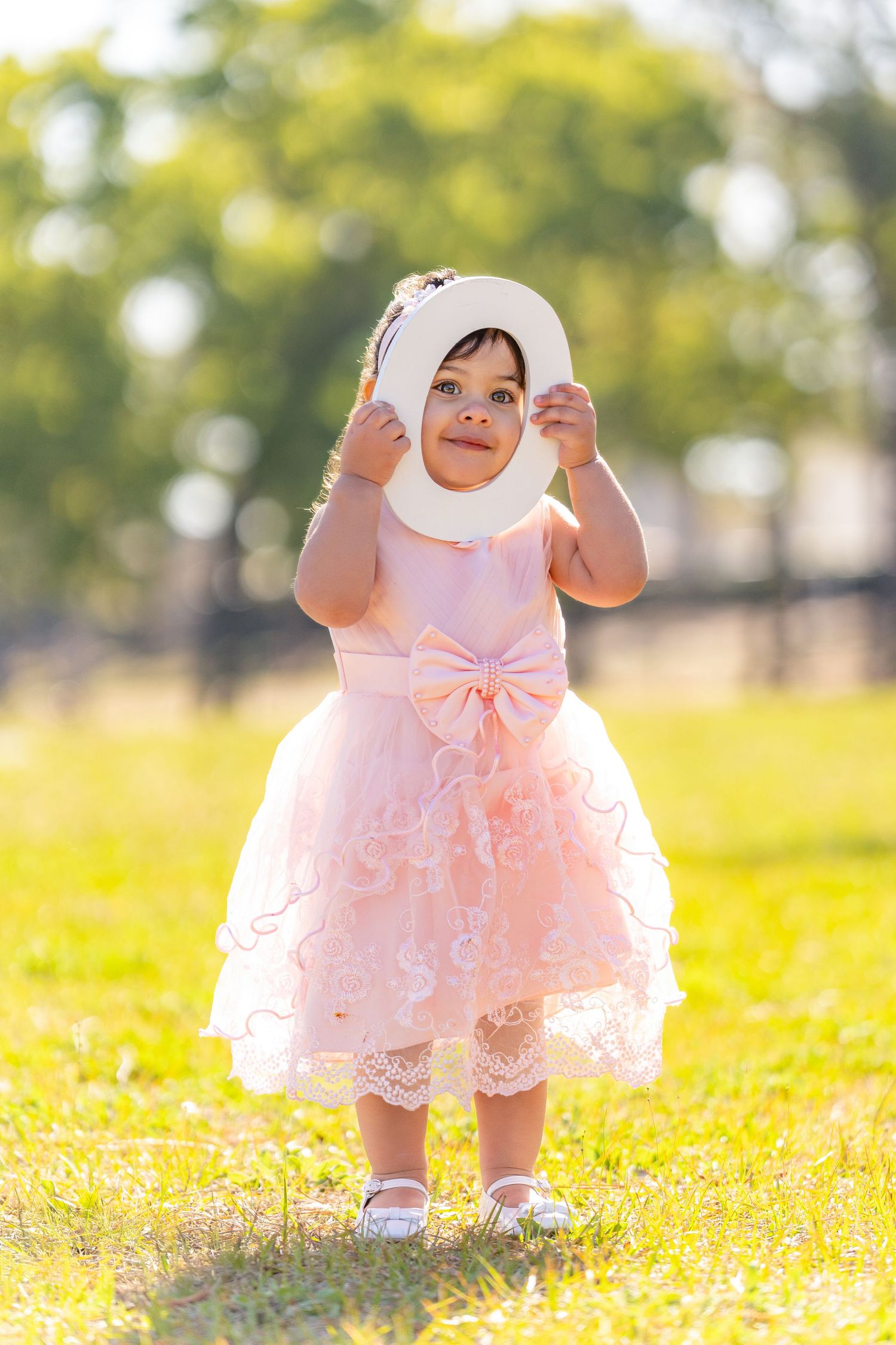 A small child wearing a pink lace dress and white hijab sits playing in a sunny field of yellow flowers.