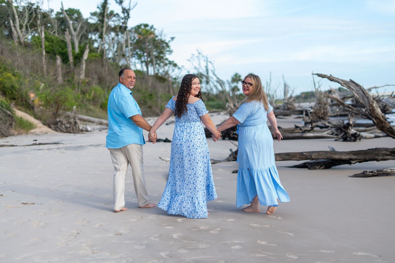 Beachgoers in flowing blue dresses walk along a scenic coastal shoreline with scattered driftwood on a bright day.