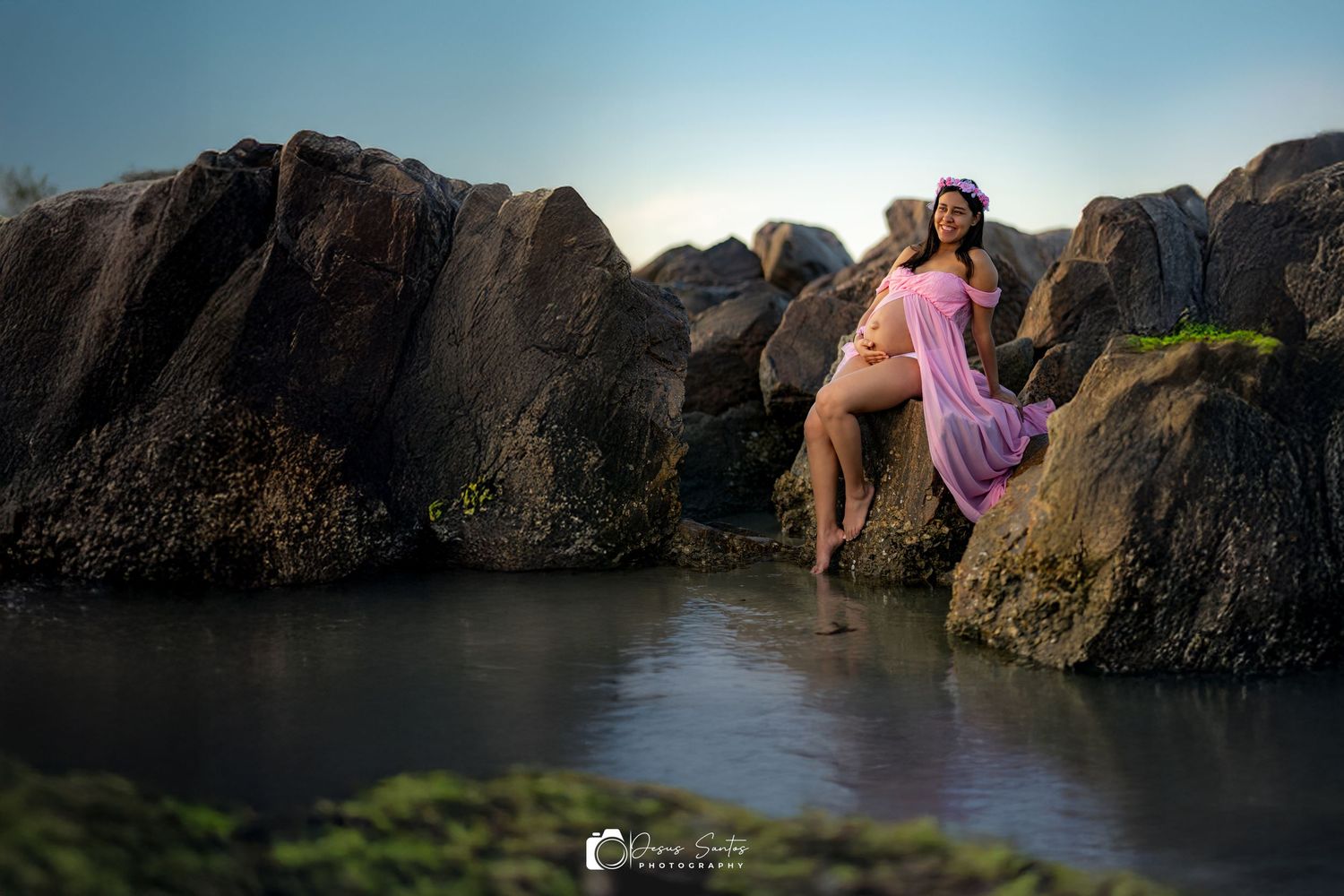 A person in a pink dress sits on dark coastal rocks near calm water at sunset creating a dreamy seaside scene.