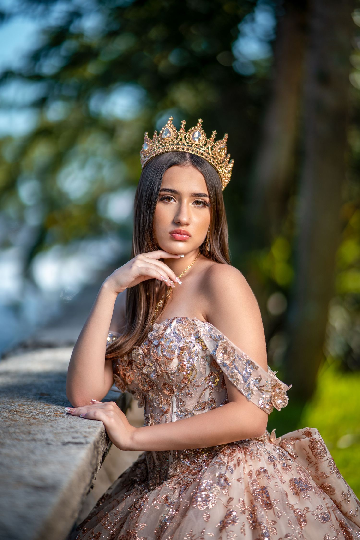 A figure in an ornate rose gold gown and crown poses outdoors against a blurred forest background.