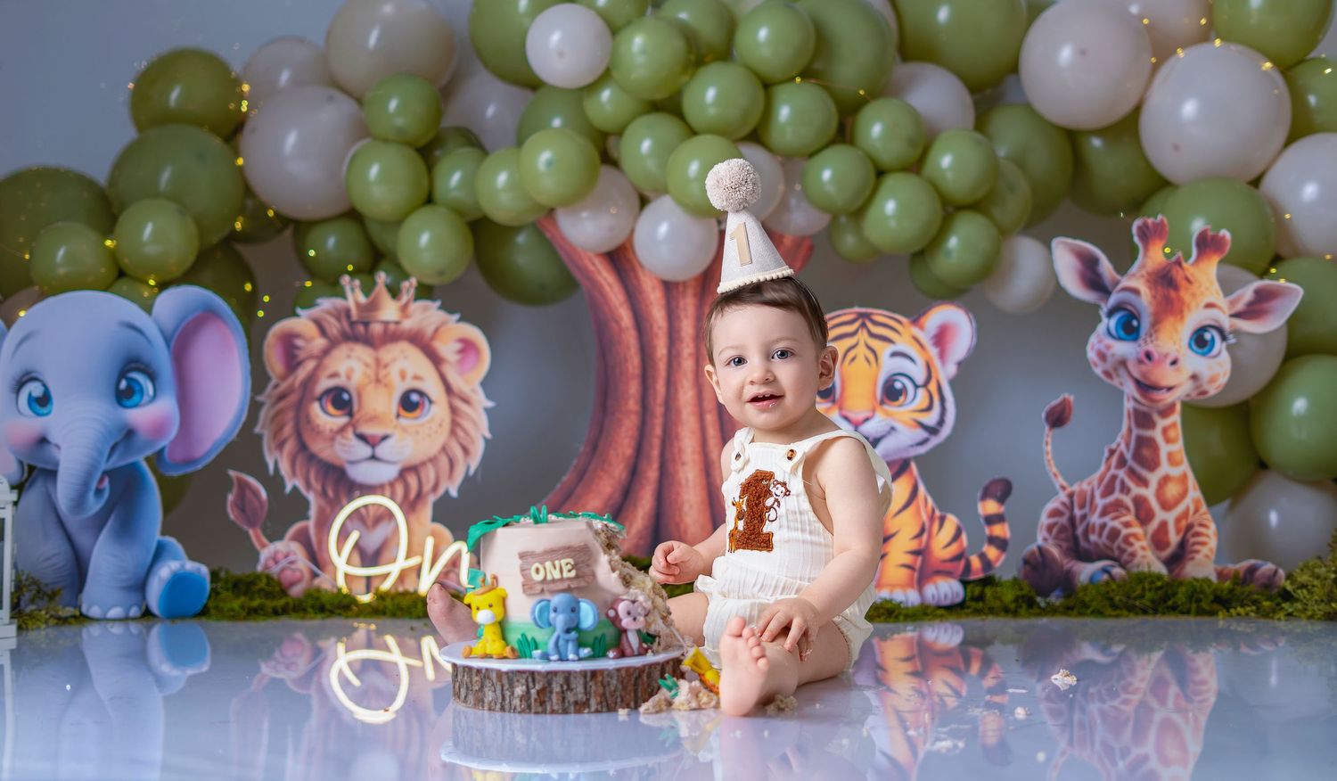 Baby in party hat celebrates first birthday with jungle animal decorations and balloon arch cake smash photo.