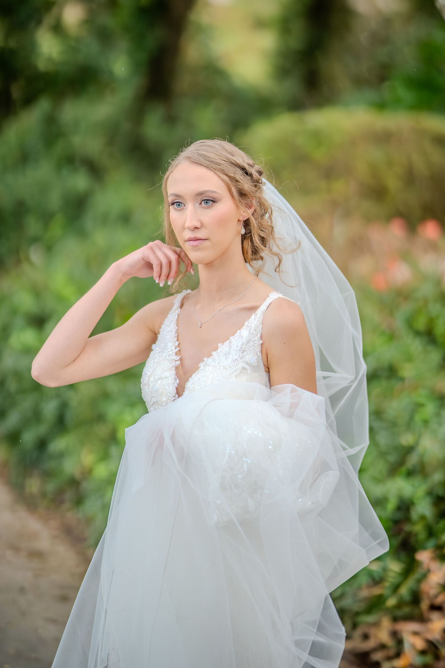 A bride in a white lace wedding dress poses gracefully in an outdoor garden setting with flowing veil and soft lighting.