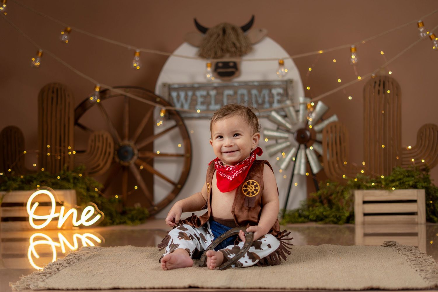 Western-themed first birthday photo setup with wagon wheel, wooden crates, string lights and rustic decorations.