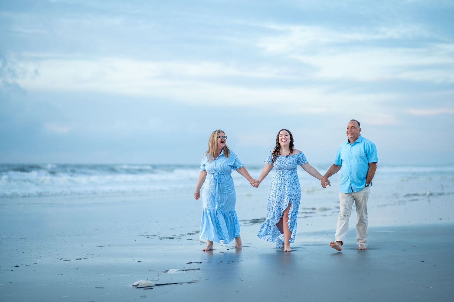 Family wearing light blue outfits walks along misty beach shore during sunset photoshoot.