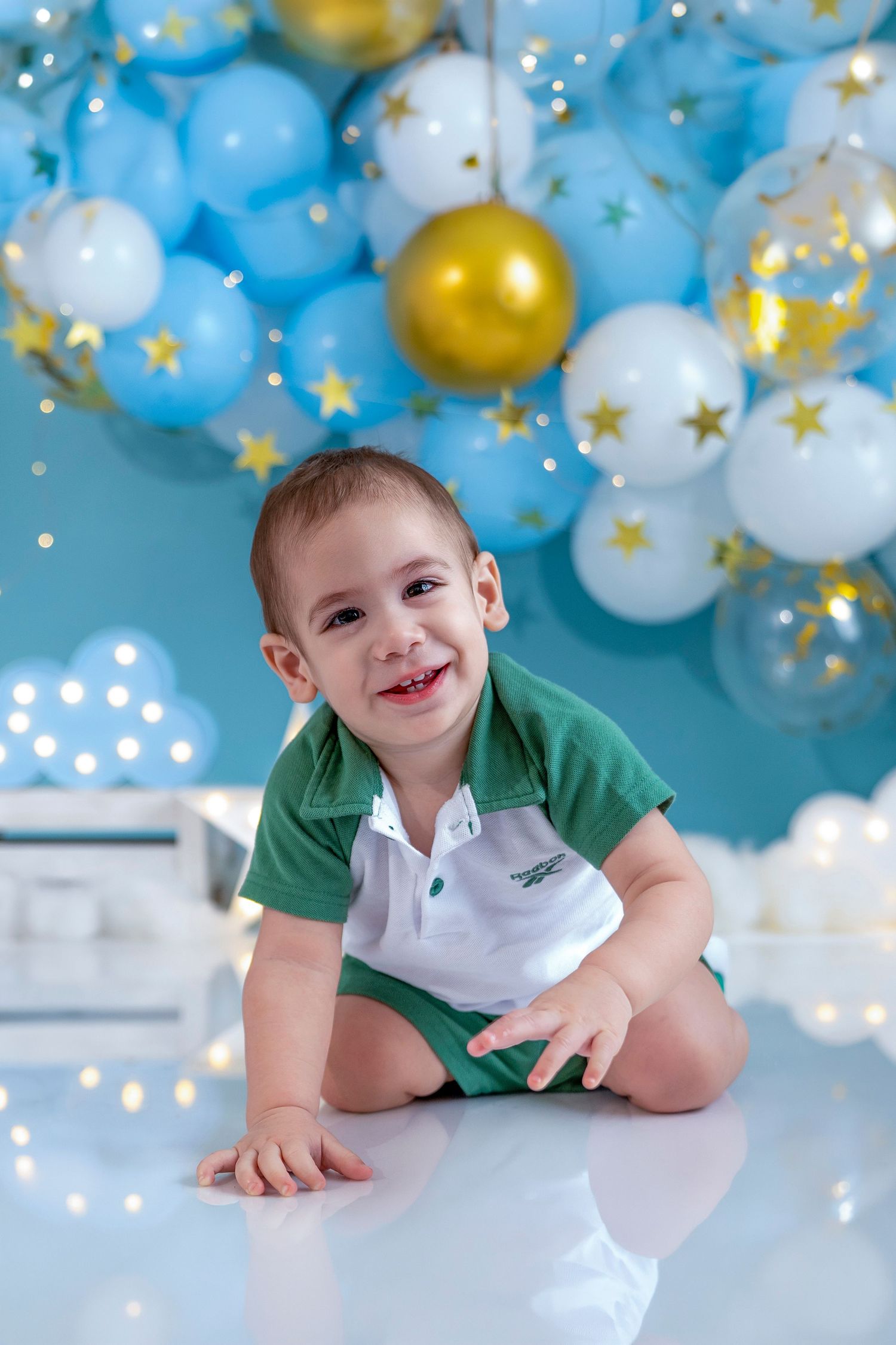 Happy toddler boy in green outfit sitting on floor with blue, white, and gold star balloons at birthday party.