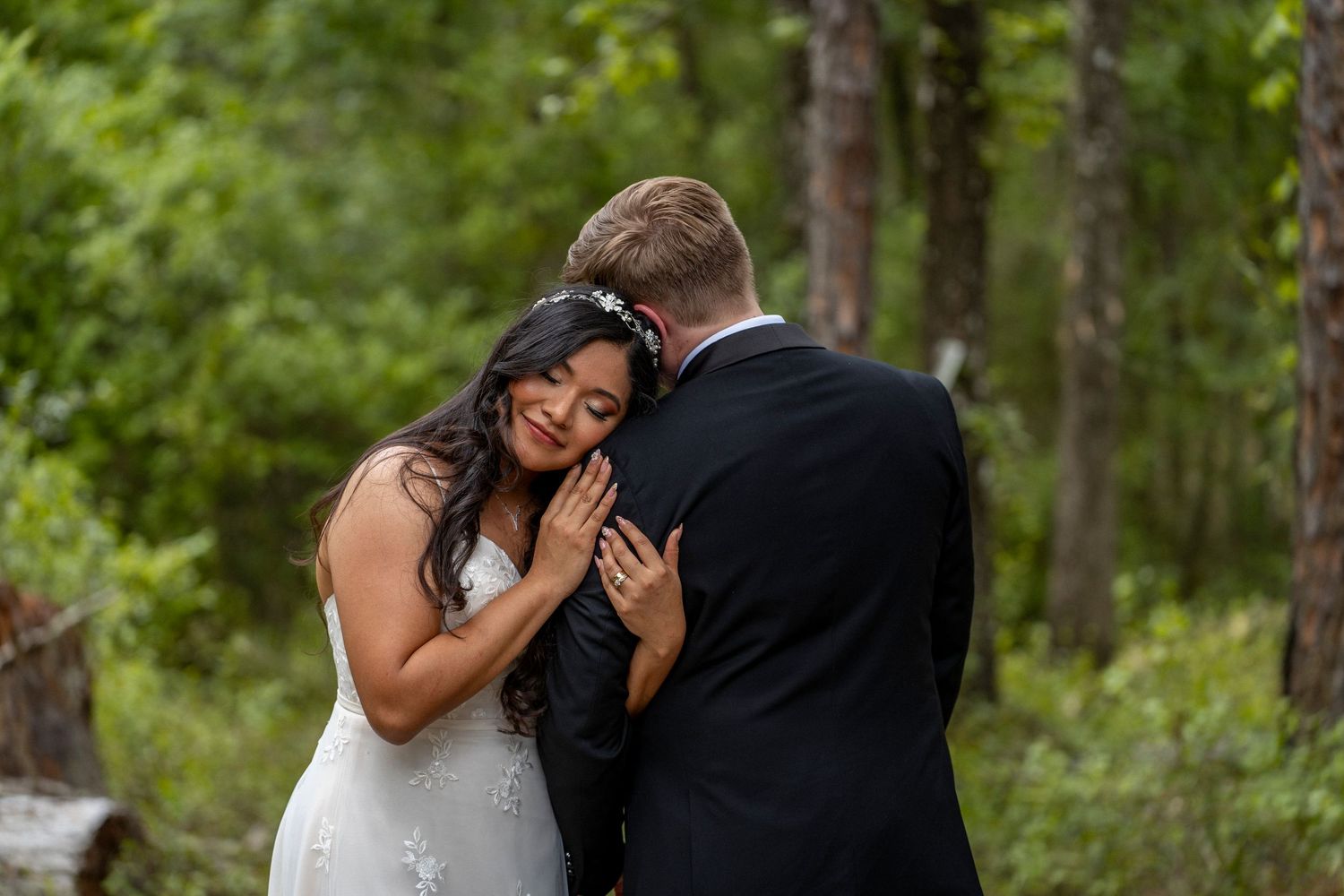 Bride in white lace gown embracing groom in black suit during outdoor forest wedding ceremony.