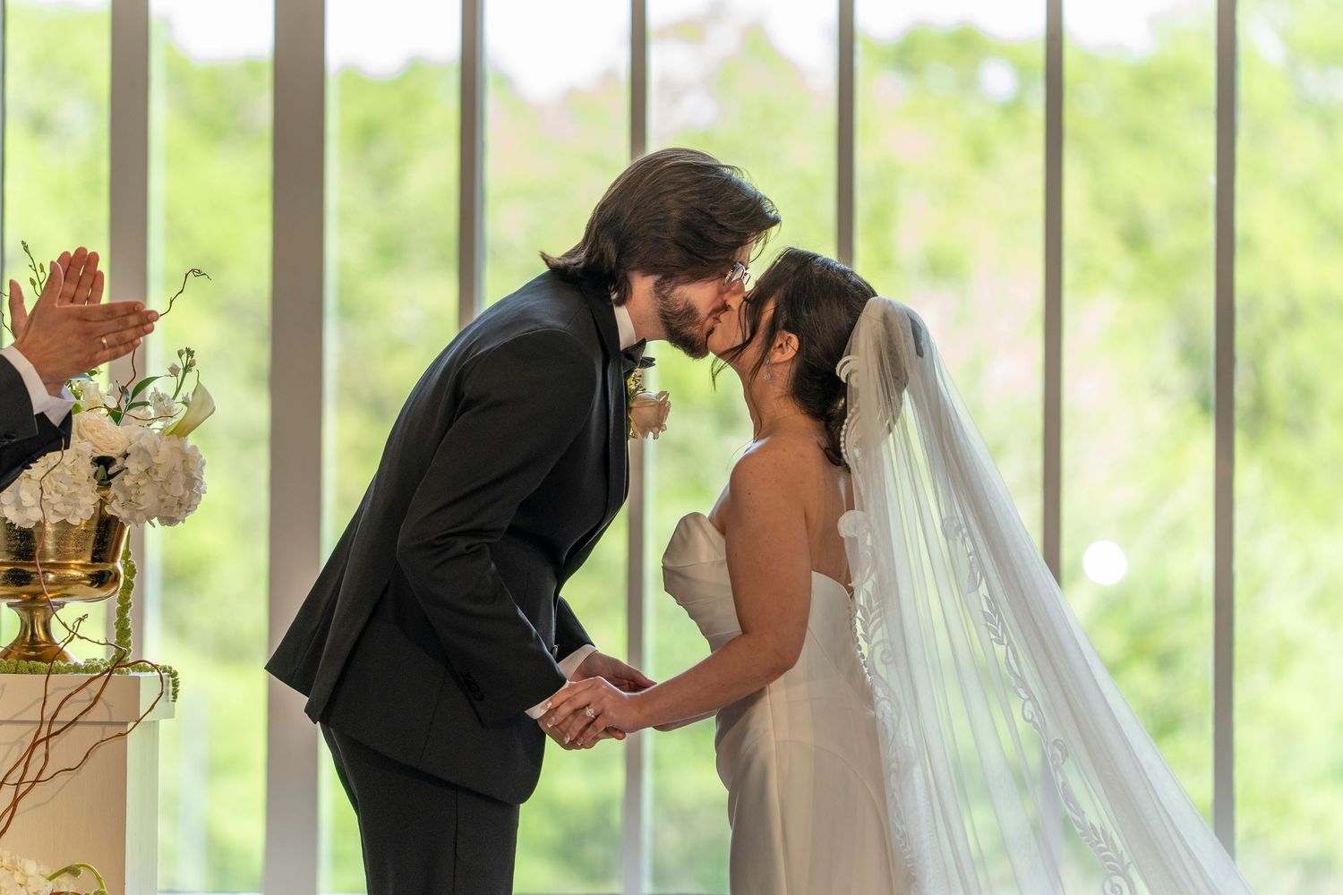 Bride and groom share first kiss at wedding altar with lush green windows in background.