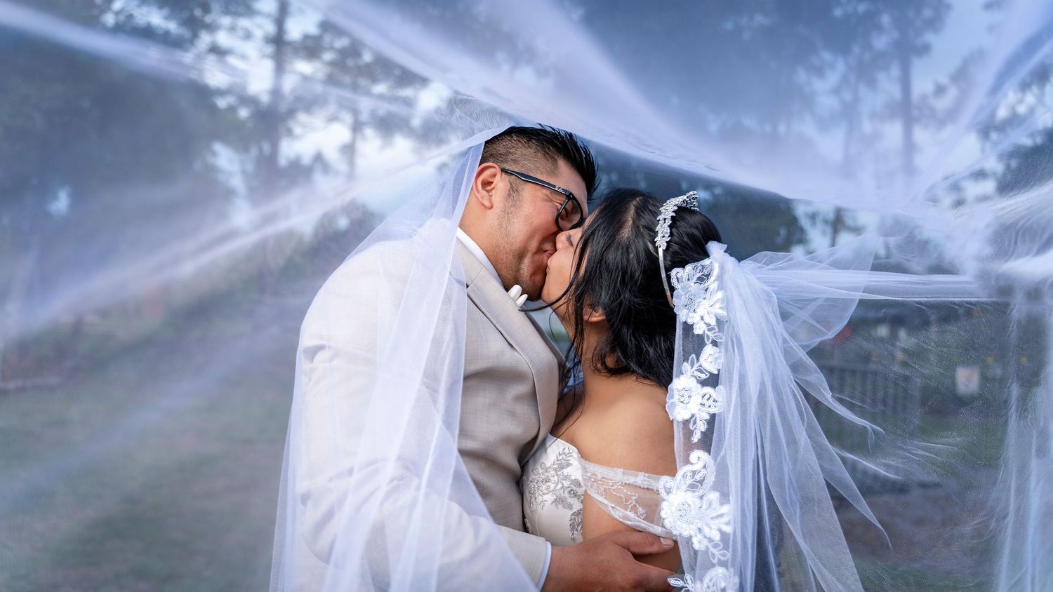 A bride and groom share a romantic kiss under a flowing white veil at their outdoor wedding ceremony.