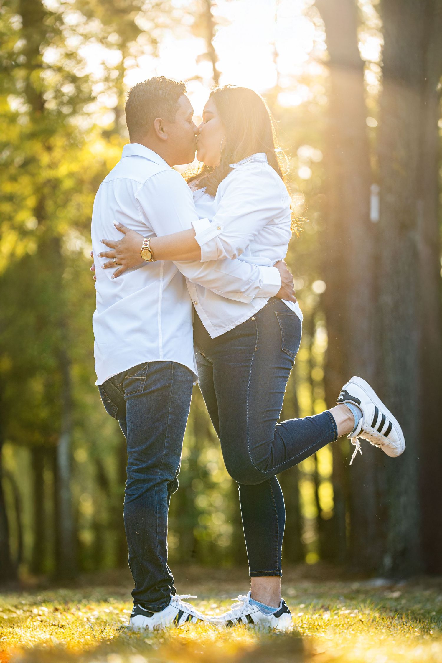 A romantic outdoor engagement photo shoot at sunset showing a couple embracing in a wooded park setting.