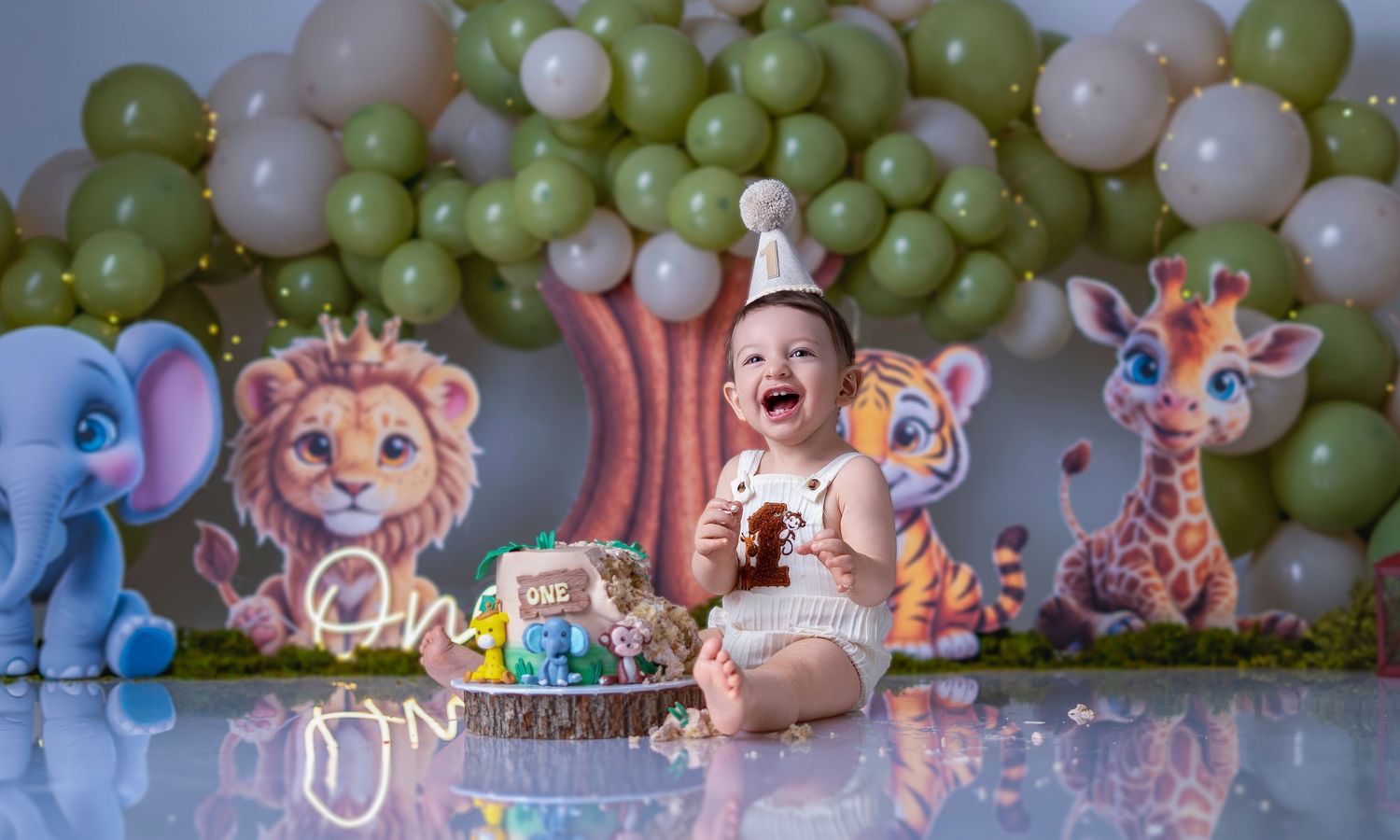 Baby in party hat smashes first birthday cake surrounded by jungle animal decorations and green balloon backdrop.