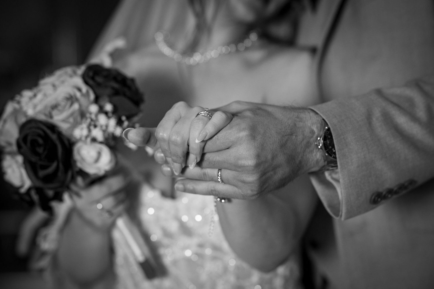 Black and white photo of bride and groom exchanging rings, holding hands with a floral bouquet nearby.