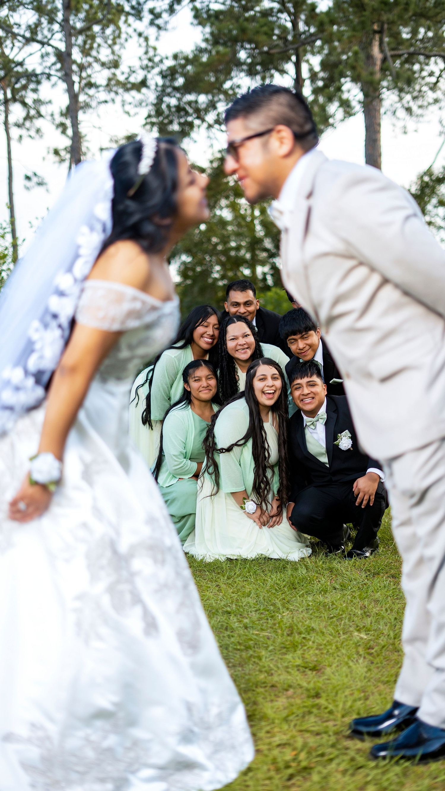 Bride and groom share a romantic moment while smiling wedding party poses on grass among trees.