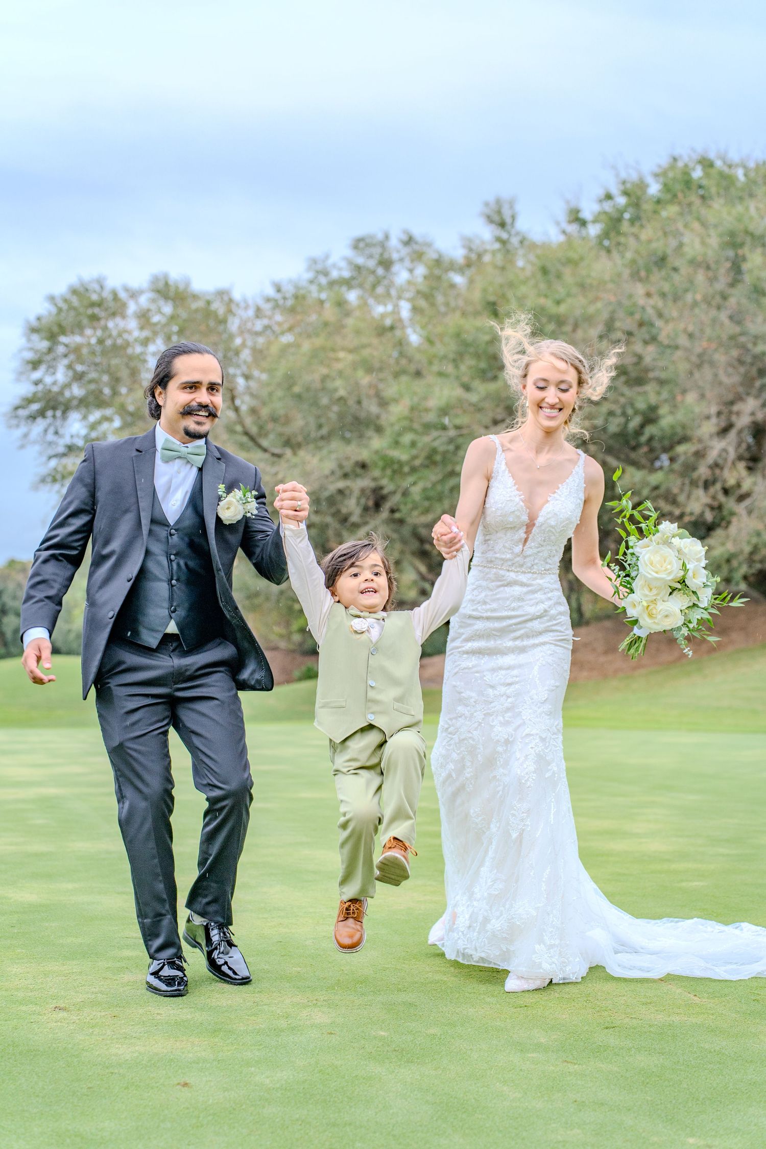 Wedding couple and child playfully posing and running together on a lush green lawn during outdoor celebration.