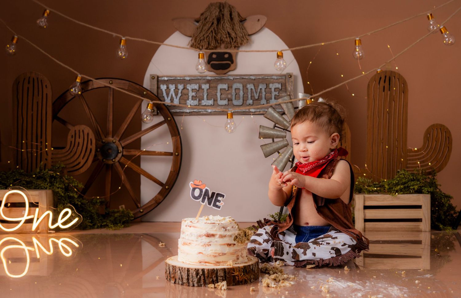 Candid moment at first birthday celebration with cake and western-themed decor including wagon wheel and lights.