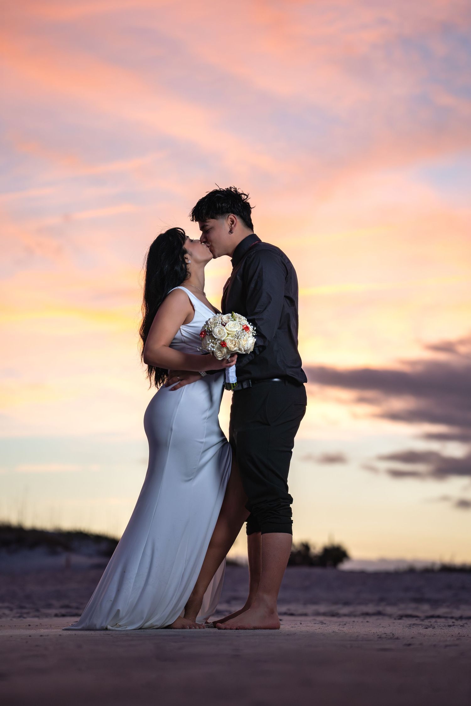 A romantic silhouette of a couple sharing a kiss at sunset on the beach with the bride holding a white bouquet.