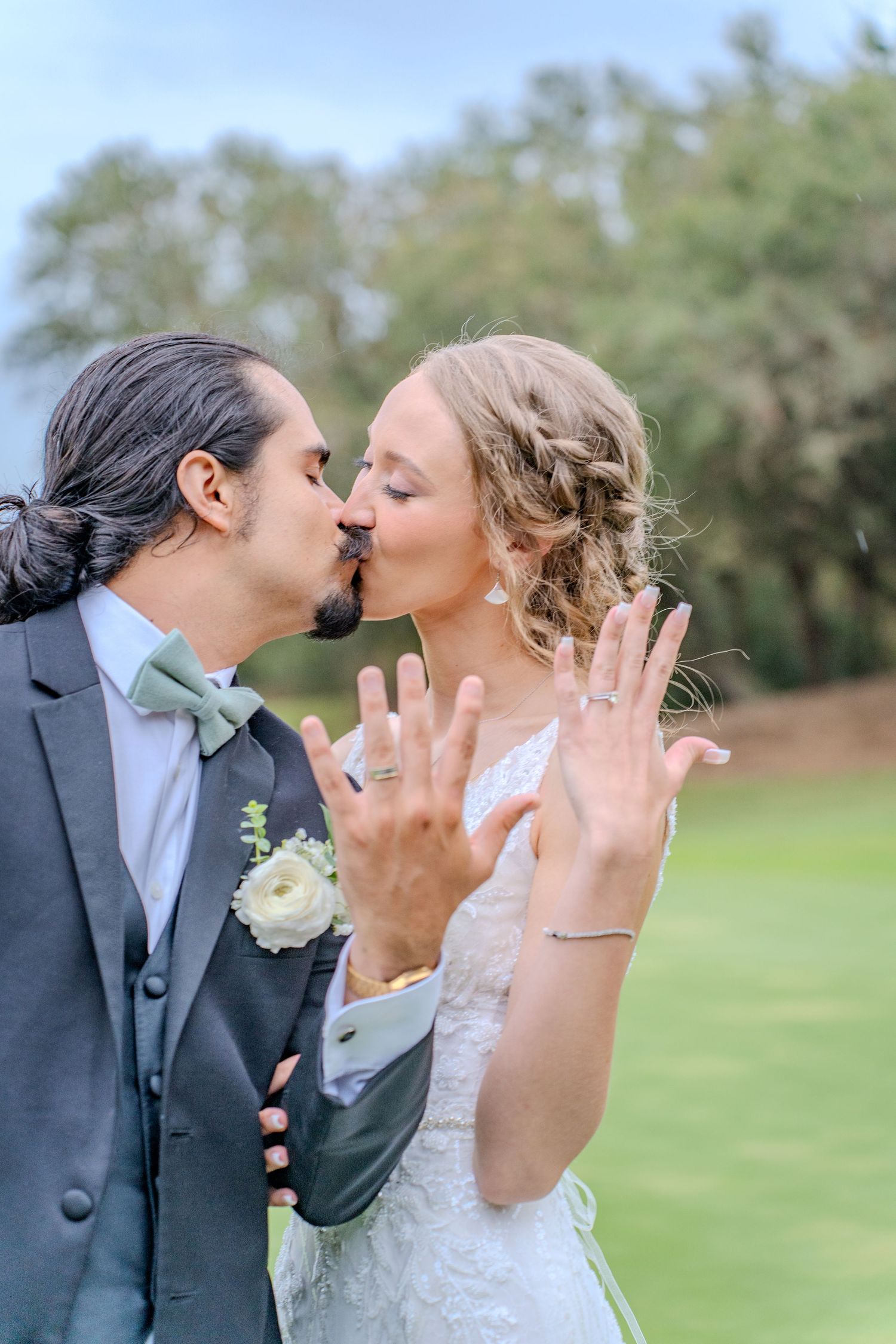 Romantic newlyweds share tender kisses at their outdoor wedding celebration on a grassy field with trees in background.