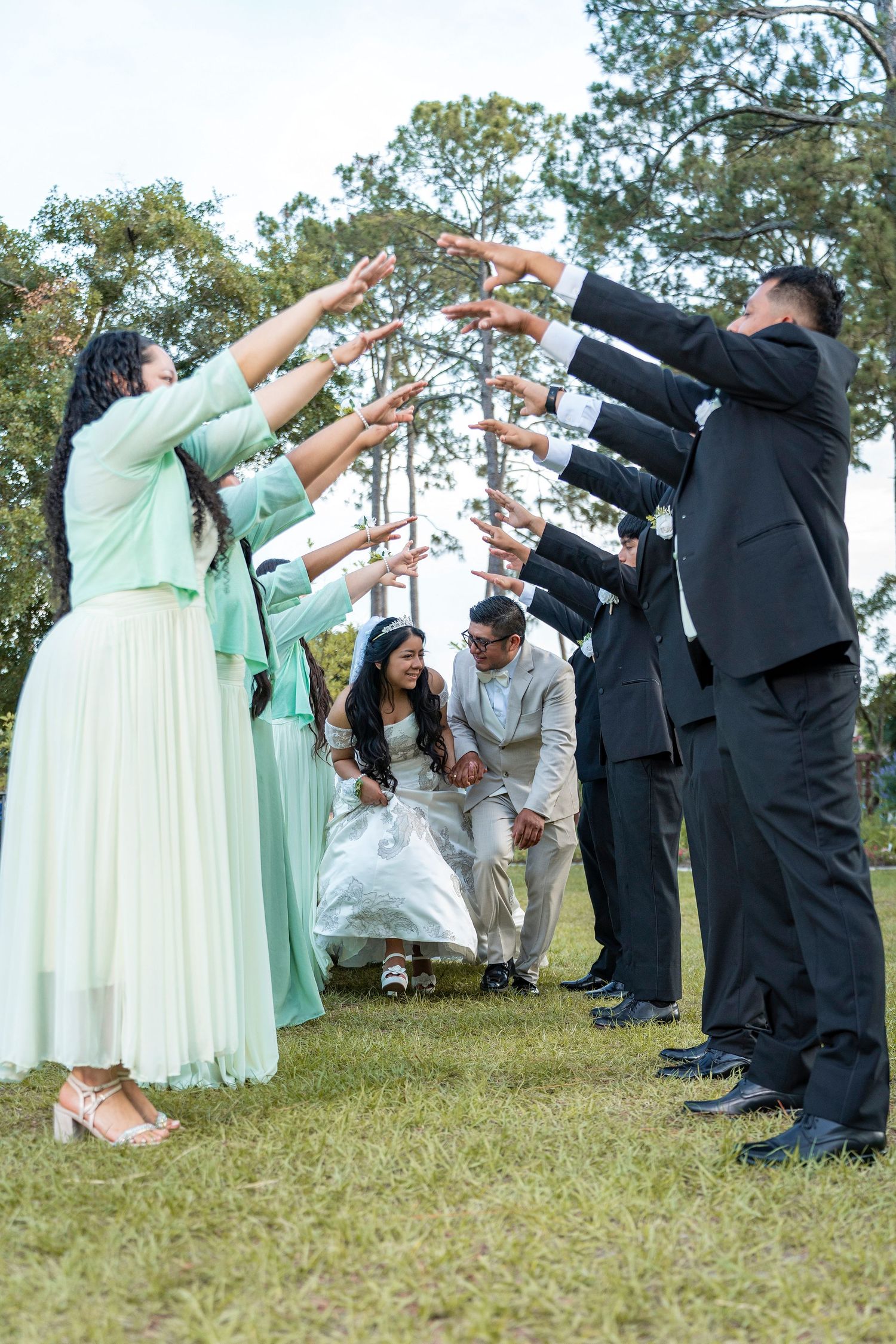Wedding party forms an arch with arms as bride and groom walk beneath during outdoor ceremony.