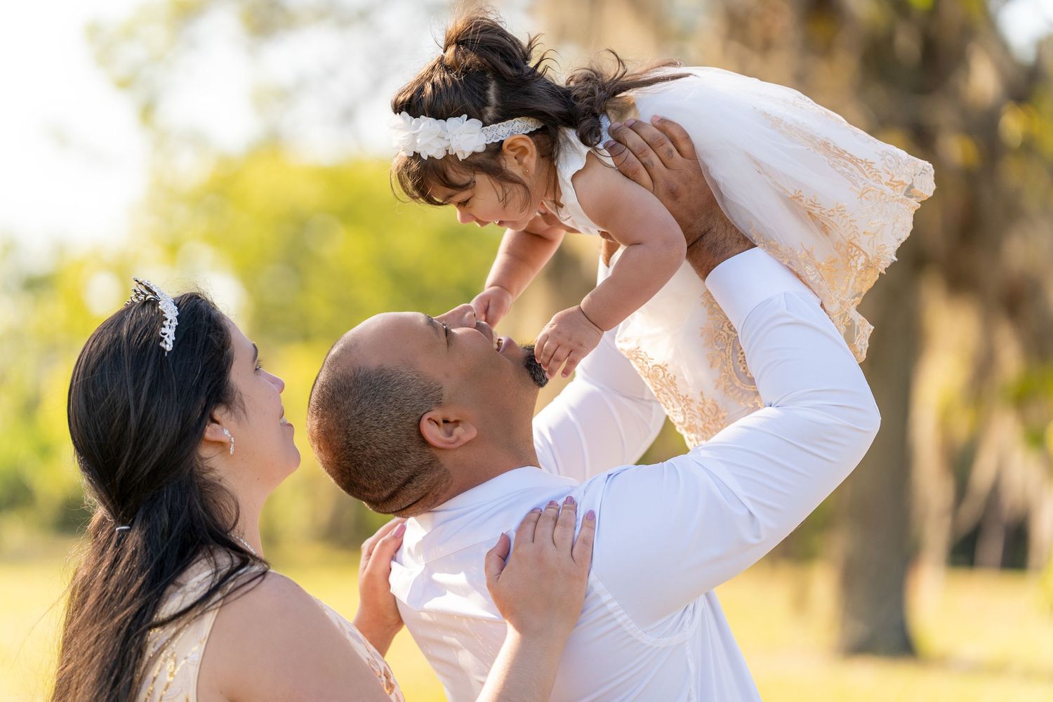 A couple in white clothing dances romantically in a sun-drenched outdoor setting with trees in the background.