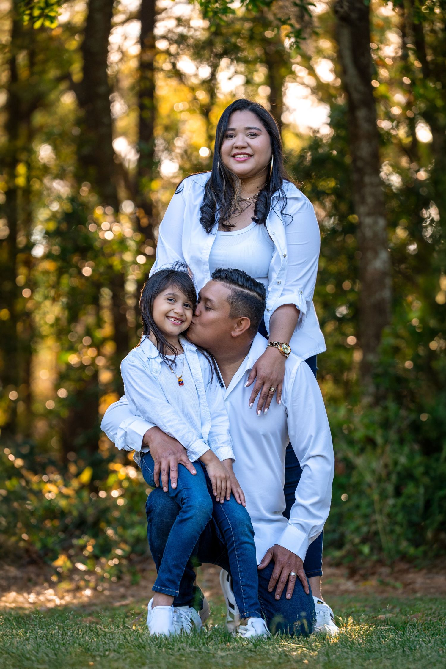 A family wearing matching white shirts and blue jeans poses together outdoors during golden hour in a park setting.