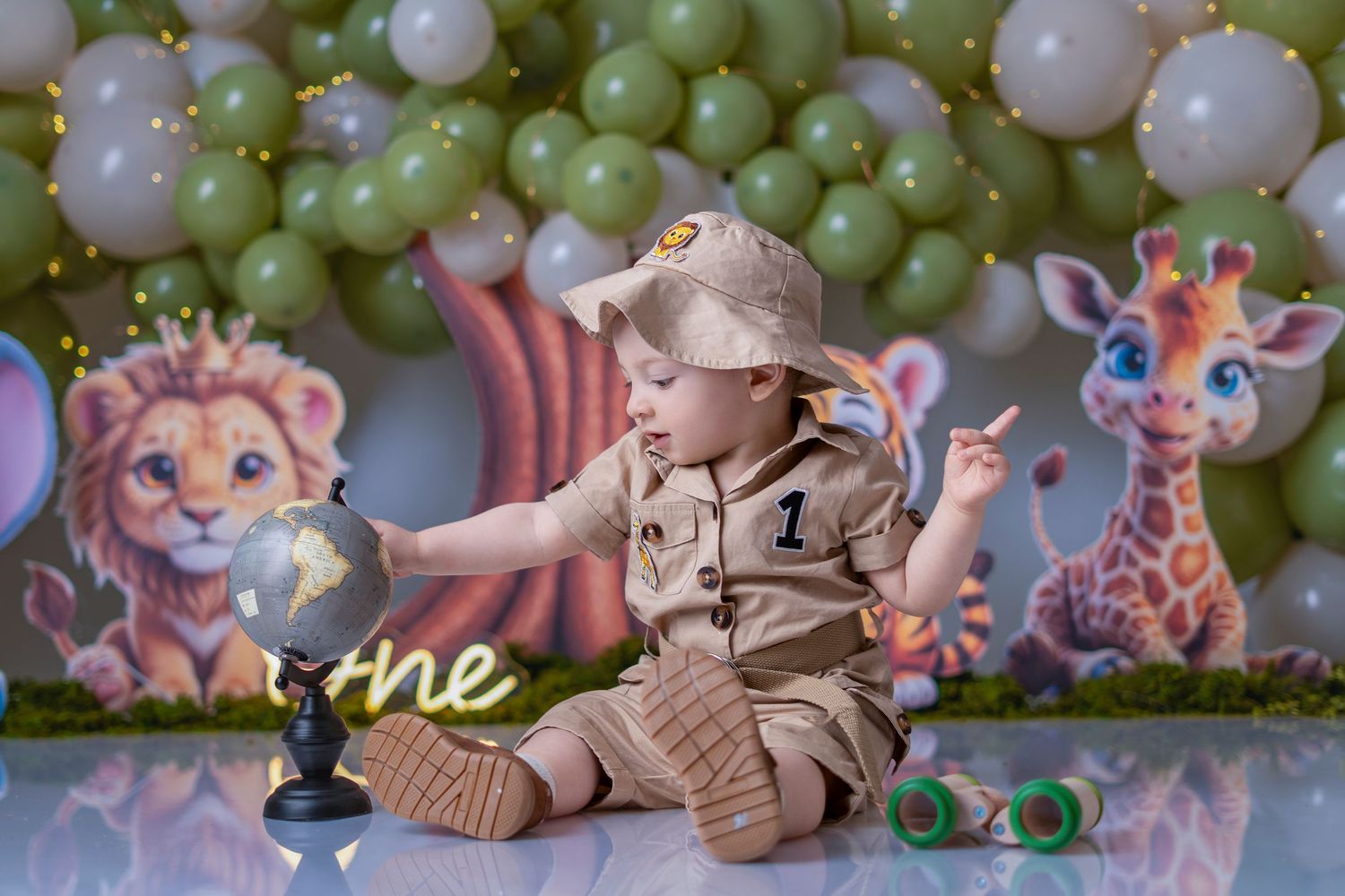 Baby in safari outfit sits with globe at jungle-themed first birthday party with balloon backdrop and animal decorations.
