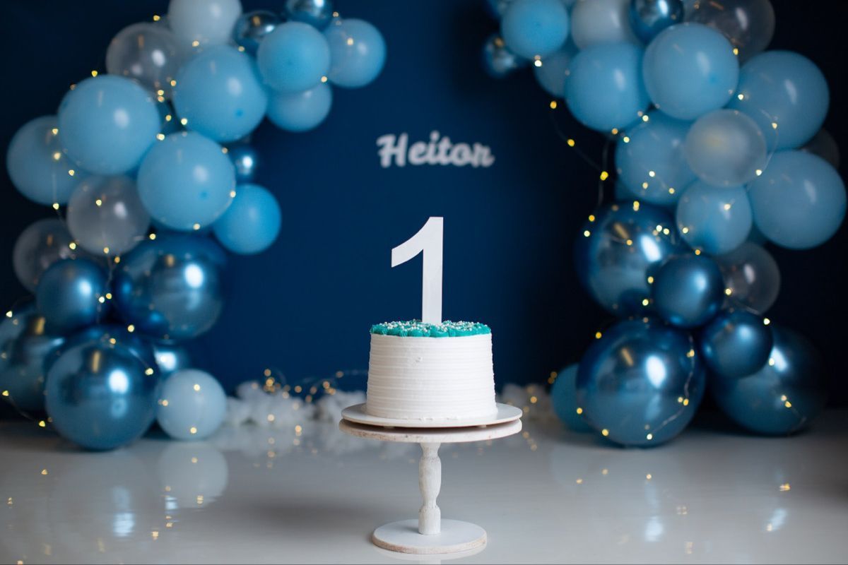 Elegant white cake on a pedestal stand with blue balloon arch decorations and sparkly lighting effects in the background.