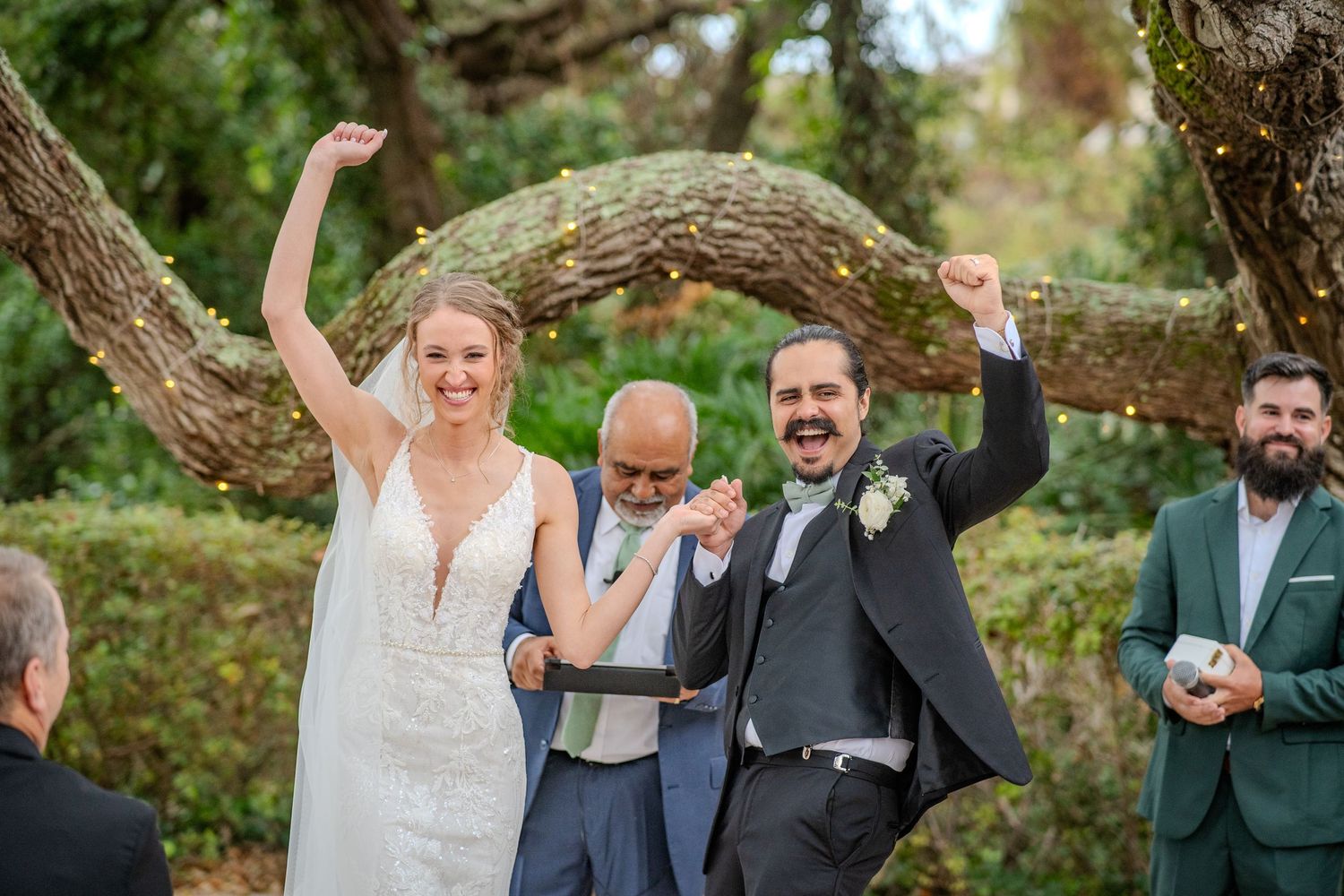 A group celebrates joyfully with raised arms during an outdoor gathering in a lush garden setting.