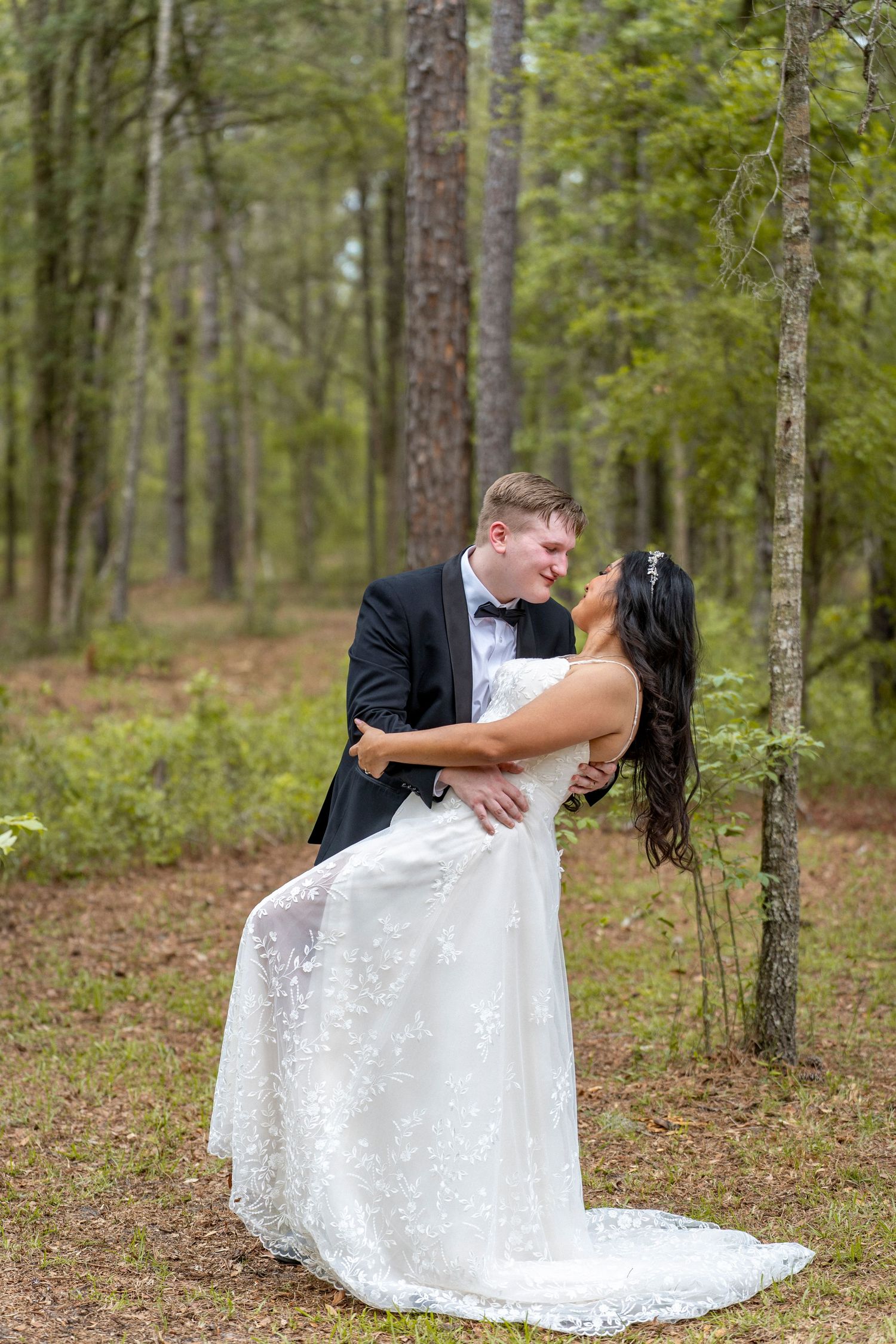 Bride and groom sharing a romantic dip kiss in a lush pine forest, bride in white lace gown, groom in black tuxedo.
