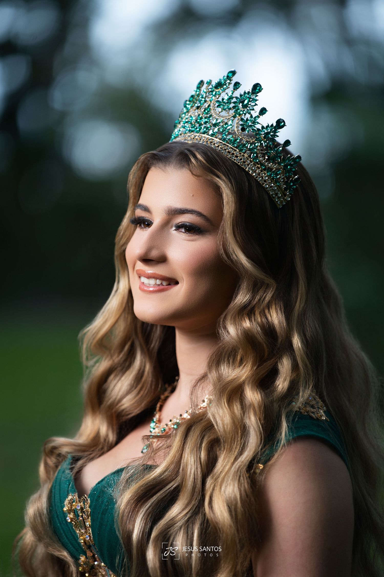 Young woman wearing an emerald green crown and matching gown, smiling outdoors with lush greenery background.