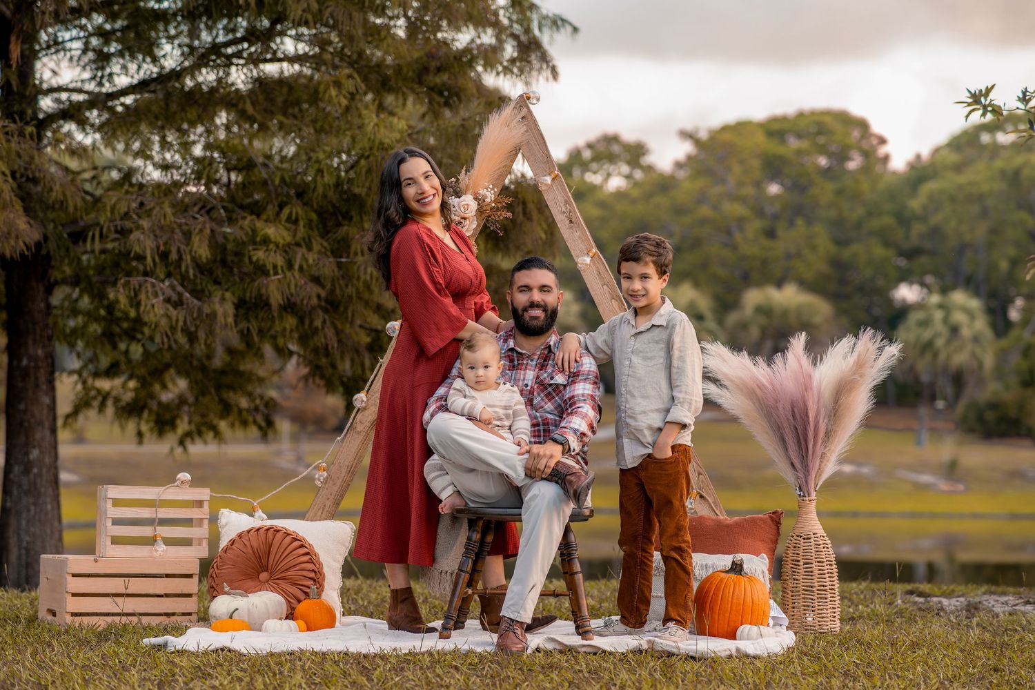 A family enjoys an autumn outdoor photo session with pumpkins and blankets in a rustic park setting at sunset.