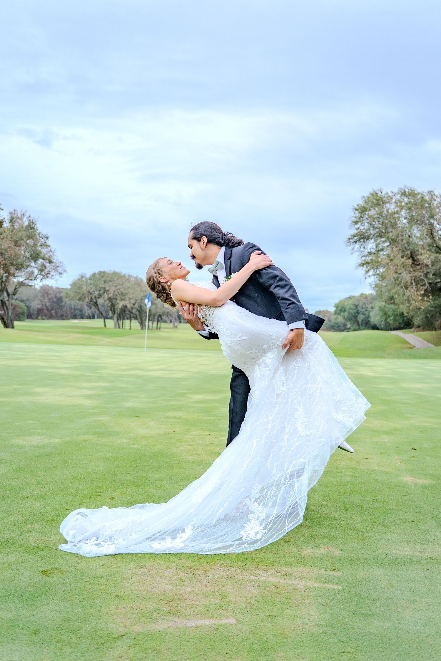 Dramatic dip kiss sequence on a golf course with flowing white wedding dress against cloudy sky.