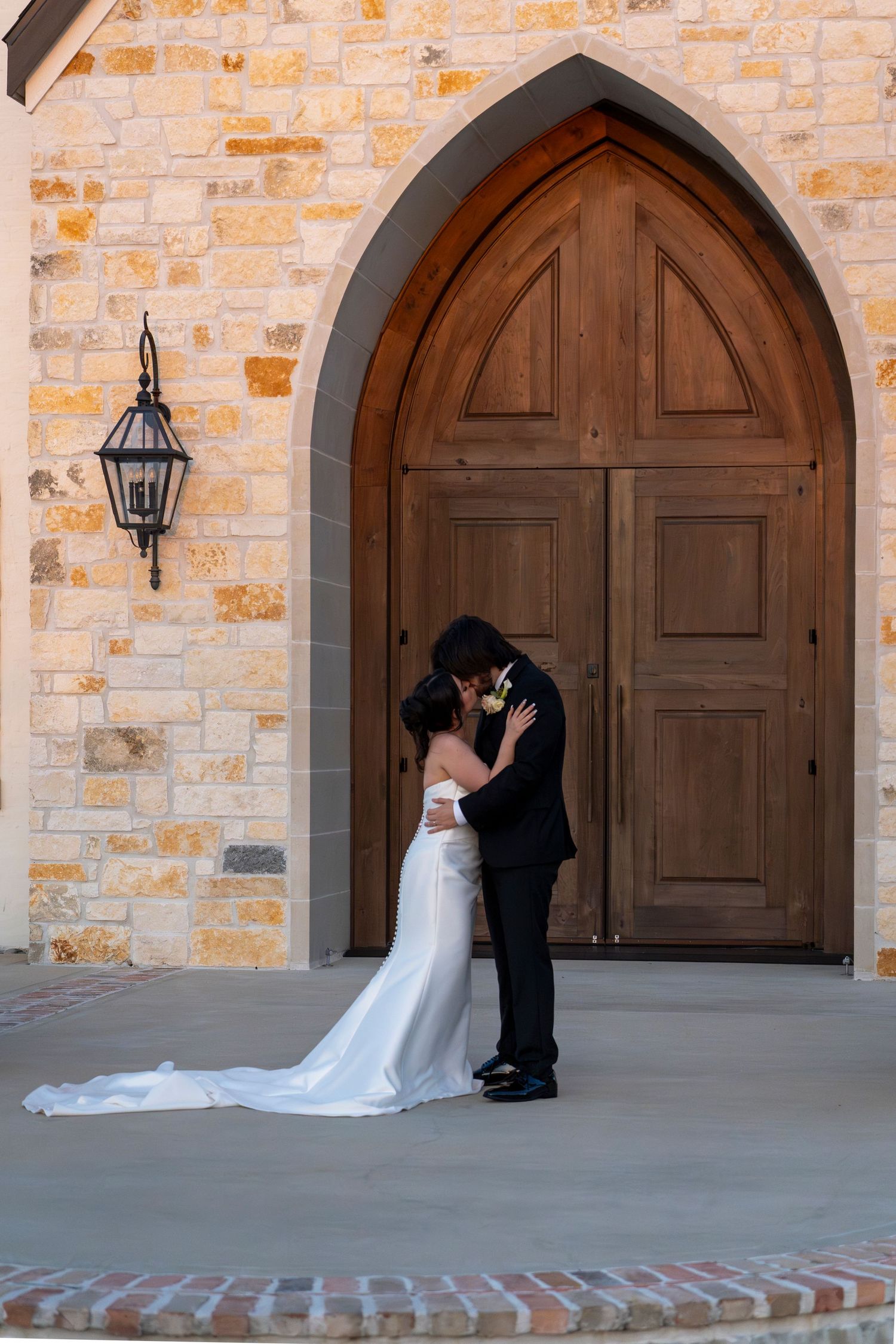 Bride and groom kiss in front of grand wooden arched doors of stone wedding chapel.