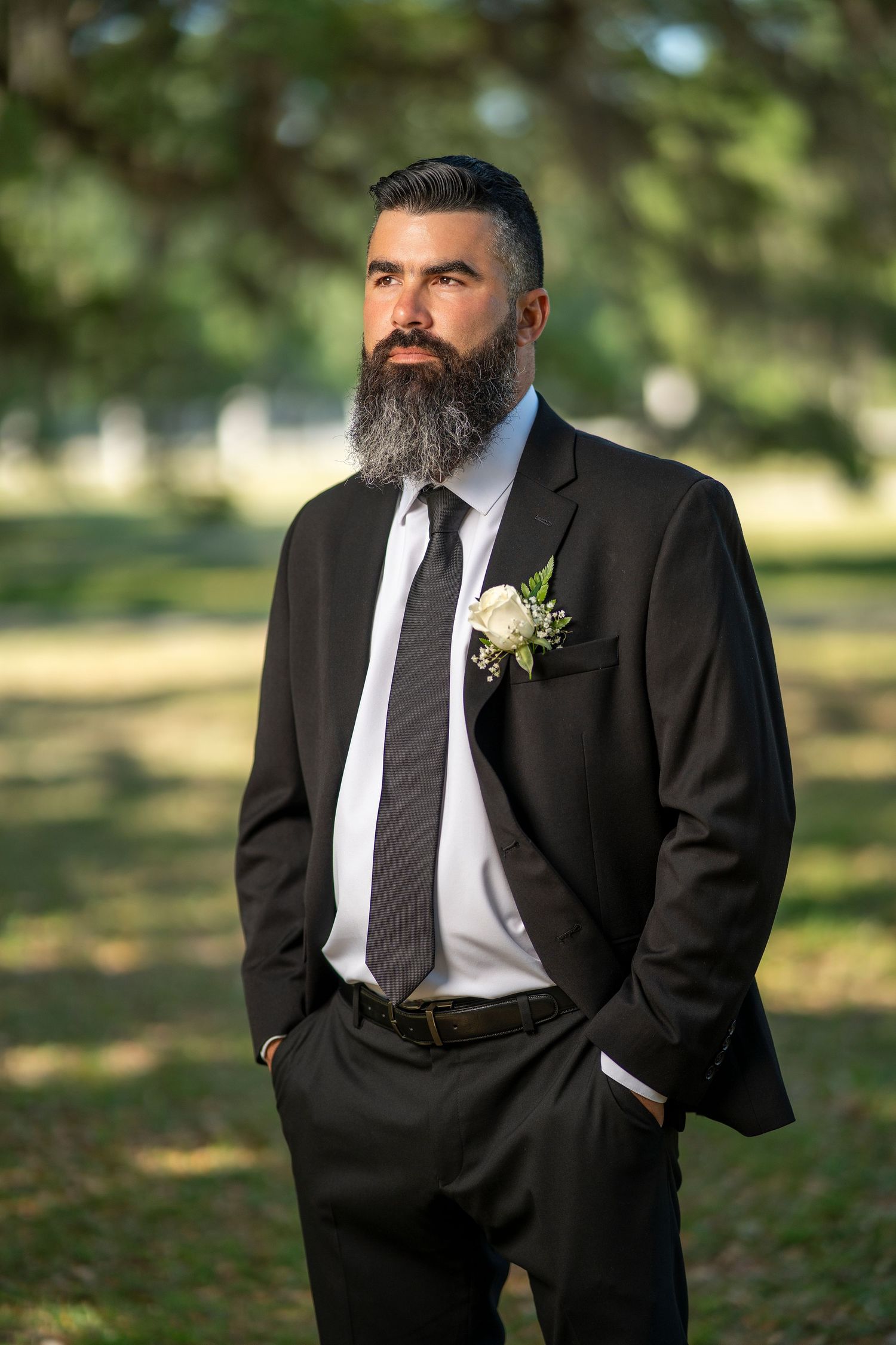 Bearded groom in black suit with white boutonniere stands outdoors in park setting for wedding portrait.