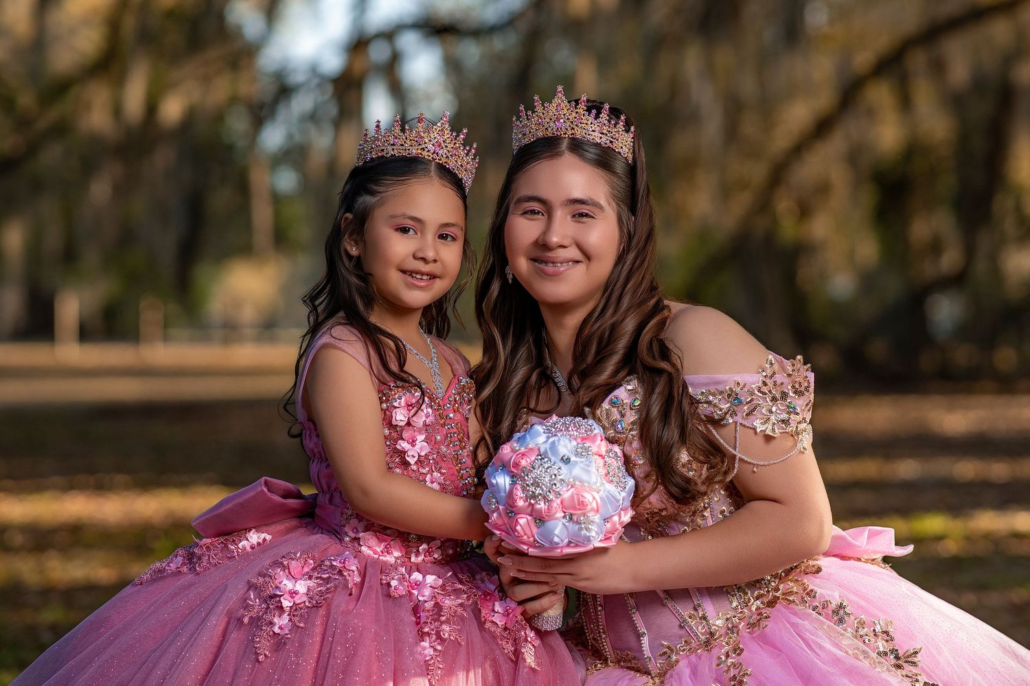 Two girls in pink ball gowns and crowns pose outdoors for a quinceañera photo session.