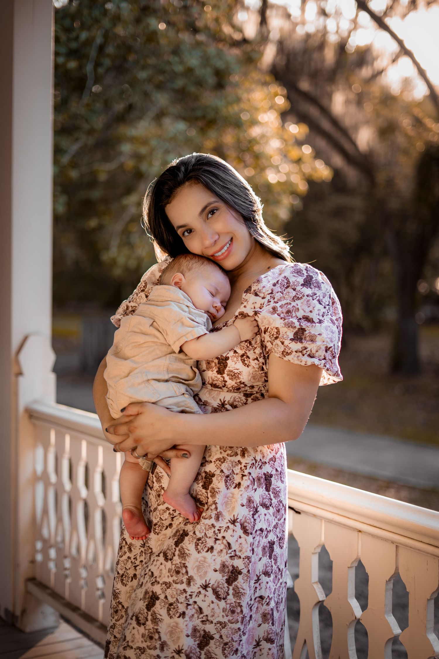 A woman in a floral dress holds and cuddles with a small cat on a front porch during golden hour.