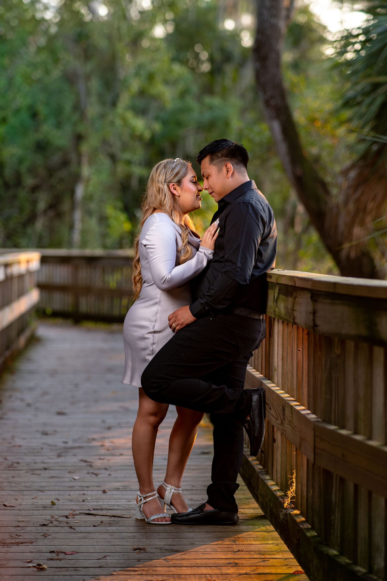A couple shares an intimate moment on a wooden boardwalk path surrounded by lush greenery at sunset.